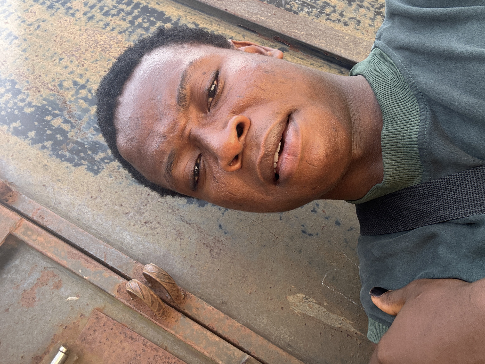 The image depicts a close-up view of a Black male wearing a green top. He is positioned in front of a metallic surface marked with rust. The individual's face displays visible sweat, with his head slightly tilted. His right hand is positioned near his torso, and one of his fingernails is painted black. The location appears to be an exterior environment in Unknown, Unknown.