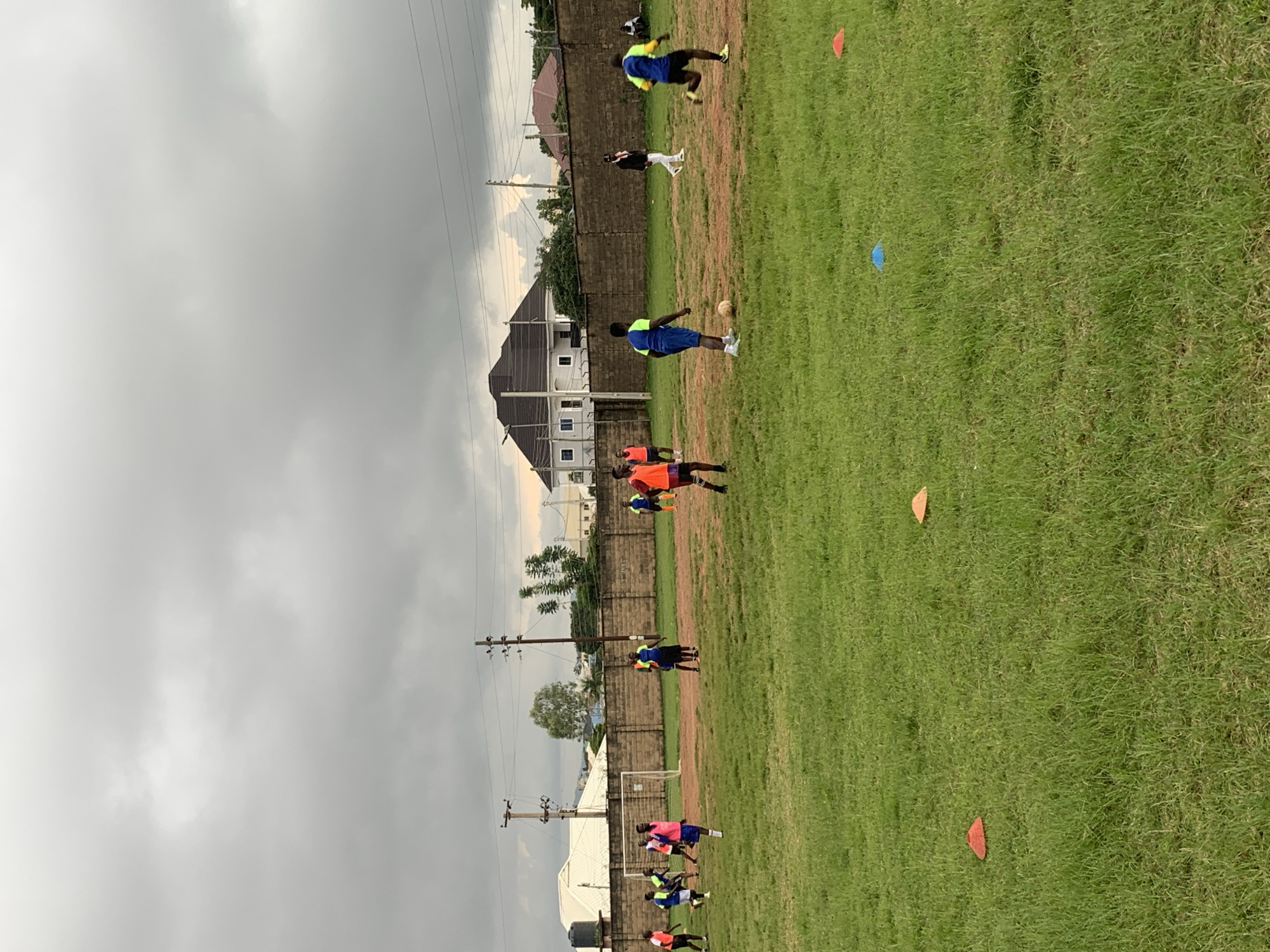 This image captures a football training session on a grassy field. Several young men, dressed in athletic wear including orange and blue jerseys, are engaged in playing soccer. Some are in motion, kicking or running with the ball, while others observe. A goalpost is visible on the left side of the frame. The background shows a brick wall, buildings, trees, and utility poles under a cloudy sky. The scene is consistent with outdoor recreational activities that might take place in a city like Jos, Nigeria, known for its football culture and numerous playing fields.