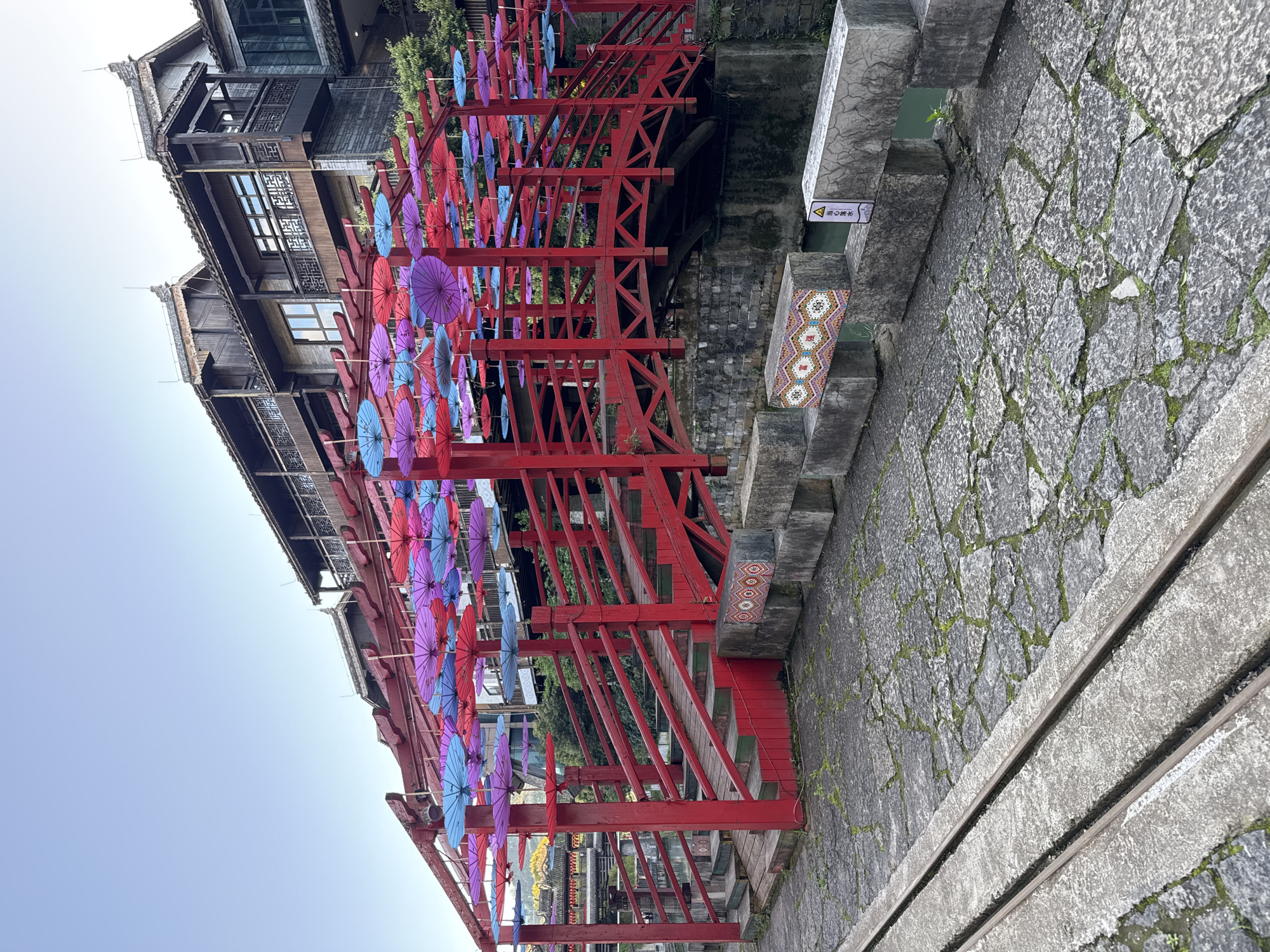 The image depicts a red-painted wooden structure, possibly a bridge or walkway, adorned with colorful umbrellas. It appears to be in Zhuoshui, China, based on the architectural style of the buildings visible in the background. The walkway is adjacent to a stone pathway, which is slightly overgrown with patches of moss. There is a warning sign near the walkway along with intricate mosaic patterns on the side of the walkway. The sky above is pale blue.