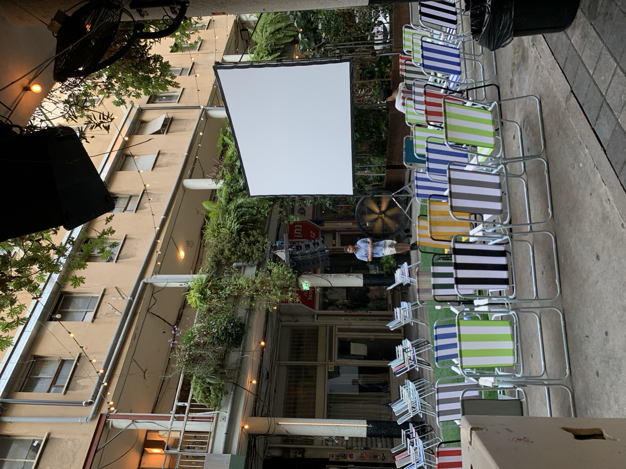 The image depicts an outdoor cinema setup in an urban courtyard in Tel Aviv, Israel. In the foreground, numerous colorful striped folding chairs are arranged in rows facing a large white projection screen. A large industrial fan is positioned to the left of the screen. A man in a light blue shirt and shorts stands in the middle ground, facing away from the camera. Behind the chairs and screen, a multi-story building with balconies and awnings is visible, adorned with hanging plants and string lights. The architecture suggests a residential or mixed-use building common in Tel Aviv. The overall scene indicates a relaxed, open-air event is about to take place or has just concluded.