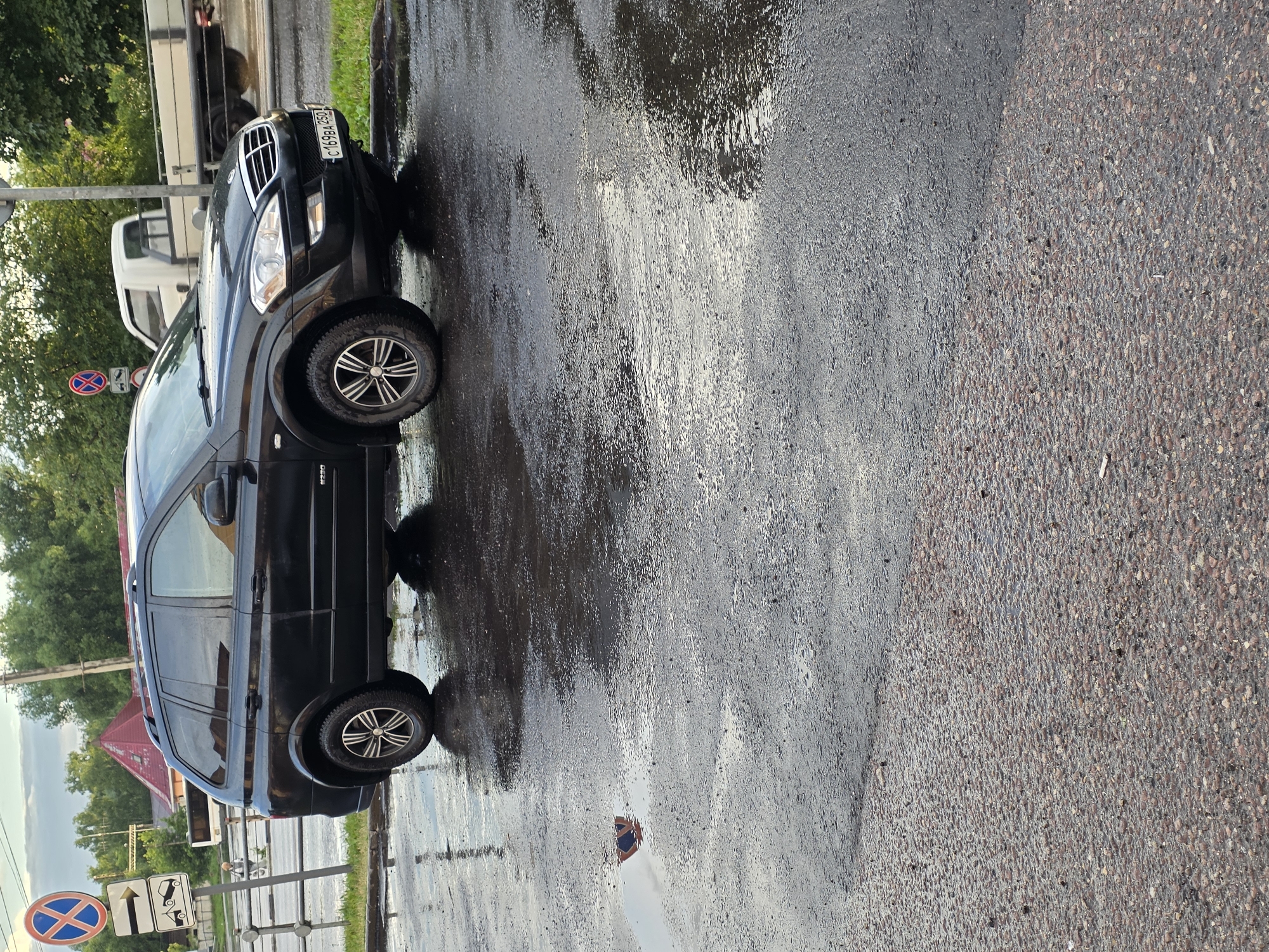A black SUV is parked on the side of a wet, asphalt road. The vehicle is angled towards the viewer, with its front end slightly turned away. The road surface is covered in puddles and reflections, indicating recent rain. In the background, there are trees, a building with a red roof, and a white truck. Traffic signs are visible on the left side of the image, including a "No Stopping" sign and an arrow indicating a direction. The sky appears cloudy. There is a reflection of a person holding a smartphone in a puddle on the road.