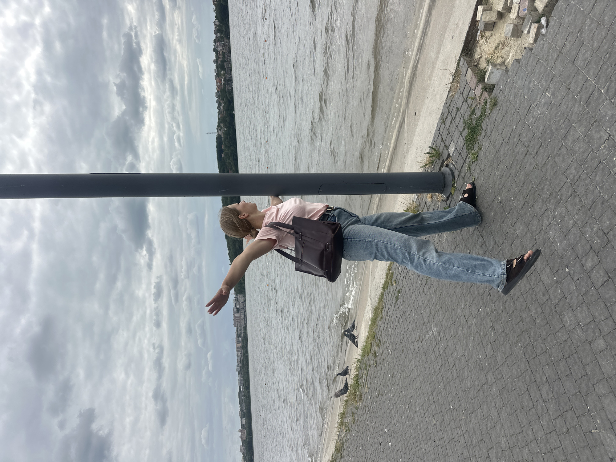 A woman stands on a paved walkway beside a body of water, with her arms outstretched as if embracing the scene. She is wearing a pink t-shirt, blue jeans, and black sandals, and carries a brown handbag. A dark pole stands to her right. In the background, a city skyline is visible across the water, with trees and buildings dotting the landscape under a cloudy sky. Several pigeons are present on the shore near the water's edge. The walkway is made of dark grey interlocking bricks, and the area to the right of the walkway shows some dislodged bricks and patches of grass. The scene appears to be located in Ternopil, Ukraine, possibly along the Ternopil Pond.