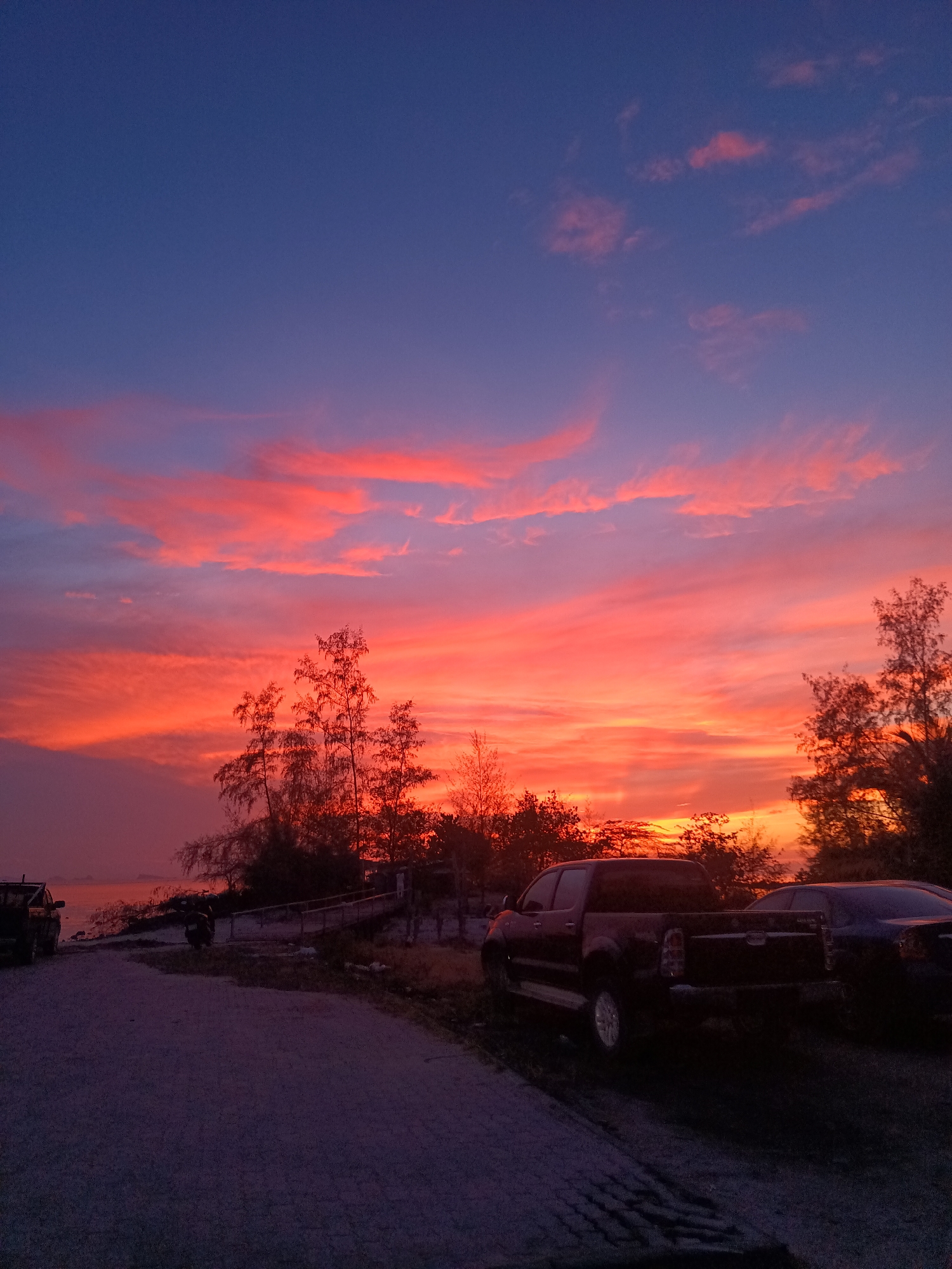 The image shows a sunset view at Baan Sri Thanu, Thailand. The sky exhibits a gradient of colors from blue to orange-red, with streaks of clouds. In the foreground, there is a paved road leading towards the water. A black pickup truck is parked on the right side of the road. Some trees and shrubs are silhouetted against the sunset. A walkway is partially visible near the trees and the water.