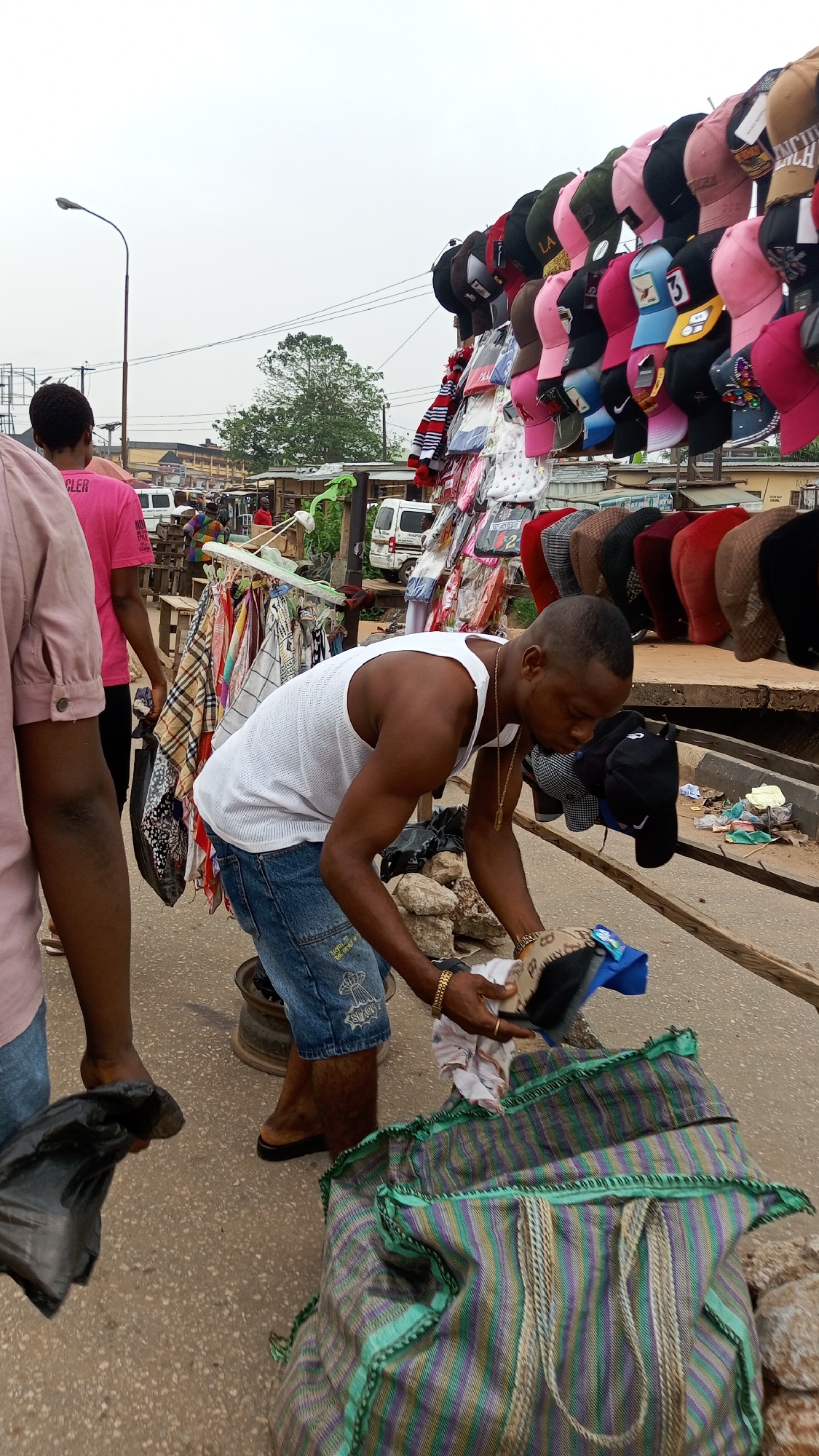 The image captures a vibrant outdoor street market scene in Onitsha, Nigeria, likely during an overcast day. The central subject is a man with short dark hair, wearing a white sleeveless tank top, blue denim shorts, a gold chain, and a gold bracelet. He is bent over, meticulously examining items—possibly caps or small accessories—from a large, striped woven bag (featuring green, purple, and beige stripes) placed on the ground in front of him. Another similar bag is partially visible behind it.
To the man's left, a person in a light pink/peach shirt and dark pants stands, holding a black object. Further back and to the left, another individual wears a bright pink t-shirt with the partial text "GLER" visible. The background reveals a bustling street with several white vans, hints of buildings, and a tall street light post.
The market stall itself is laden with merchandise. Above and to the right of the main subject, numerous baseball caps of various colors (including pink, black, red, green, blue, and brown) are displayed on racks. Some caps feature visible logos such as "LA" and a Nike swoosh. Below the caps, various smaller bags or pouches are also hanging. To the left of the central activity, another display rack holds various patterned fabrics or scarves. The ground is a mix of asphalt and dirt, with rocks and debris scattered, characteristic of an active marketplace. The text "MY STYLE" and some faint graphics are visible on the man's denim shorts.