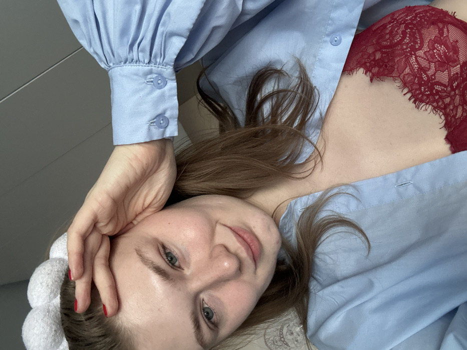 A woman poses looking at the camera while laying down. She is wearing a light blue, button-up shirt that is partially unbuttoned, revealing a red lace bra. Her head is resting on her hand, and she has a white, puffy headband on. Her hair is light brown and loose around her face.