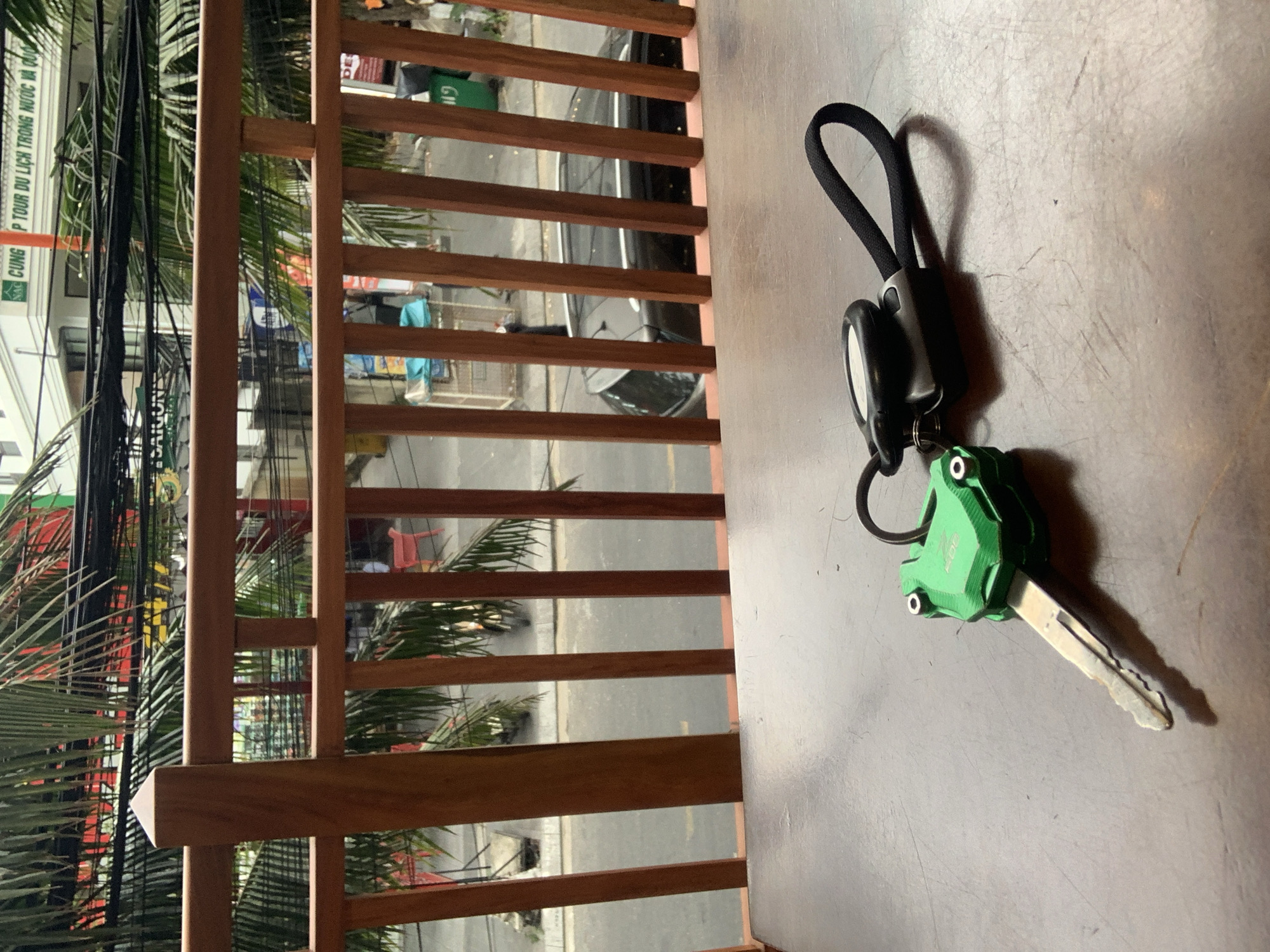 A close-up, eye-level shot taken from a high vantage point shows keys on a surface. The keys feature a black fob connected to a green, frog-shaped attachment. The scene is viewed through a wooden railing. Outside the railing, a street scene unfolds, dominated by buildings, palm trees, and an array of power lines. The street is gray, and cars are visible. A sign on a building reads “TOUR DU LỊCH TRONG NƯỚC.” The location is identified as Đà Nẵng, Vietnam. No people are visible in the image.