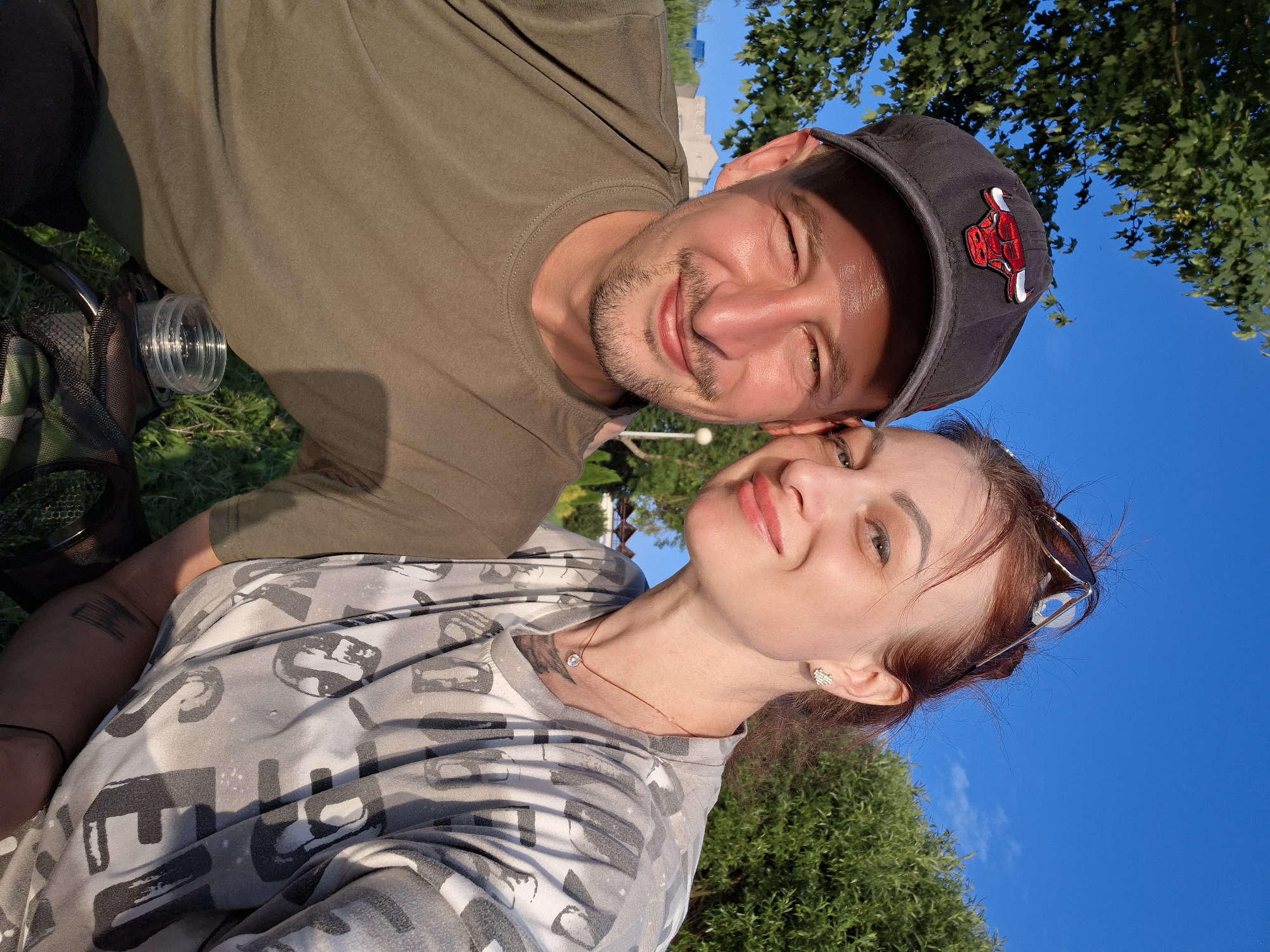 In Myadzyel, Belarus, a man and a woman are captured in a selfie outdoors. The man, wearing a dark baseball cap with a Chicago Bulls logo, smiles at the camera. He has short hair and stubble and is dressed in a green t-shirt. The woman, with red hair and sunglasses perched on her head, also smiles. She wears a grey and white camouflage t-shirt adorned with a pattern of letters and a delicate necklace with a single pendant. A small, clear container is visible in the man's lap. The background features lush green trees, a bright blue sky, and hints of buildings and outdoor lighting, suggesting a park or a recreational area. The lighting indicates it is daytime, likely late afternoon, due to the warm, golden hue cast on the subjects.