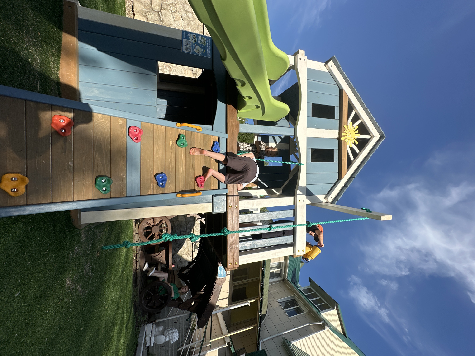 In the city of Danilovka (Altai), Russia, a sun-drenched outdoor scene unfolds featuring a multi-level wooden playground. The playground, painted in shades of blue and yellow with a green slide, is the central focus. A young boy, wearing brown shorts, is actively climbing the playground's wooden ladder with colorful climbing holds. Another child, partially obscured, stands on the playground's upper level. To the right, a yellow telescope is held by someone wearing an orange hat. In the background, a light-colored building with a green roof is visible. Below the playground, a woman in a green dress sits on a bench under a rustic awning, surrounded by decorative wagon wheels. A stone wall and a manicured lawn complete the environment, all under a bright blue sky with scattered clouds.