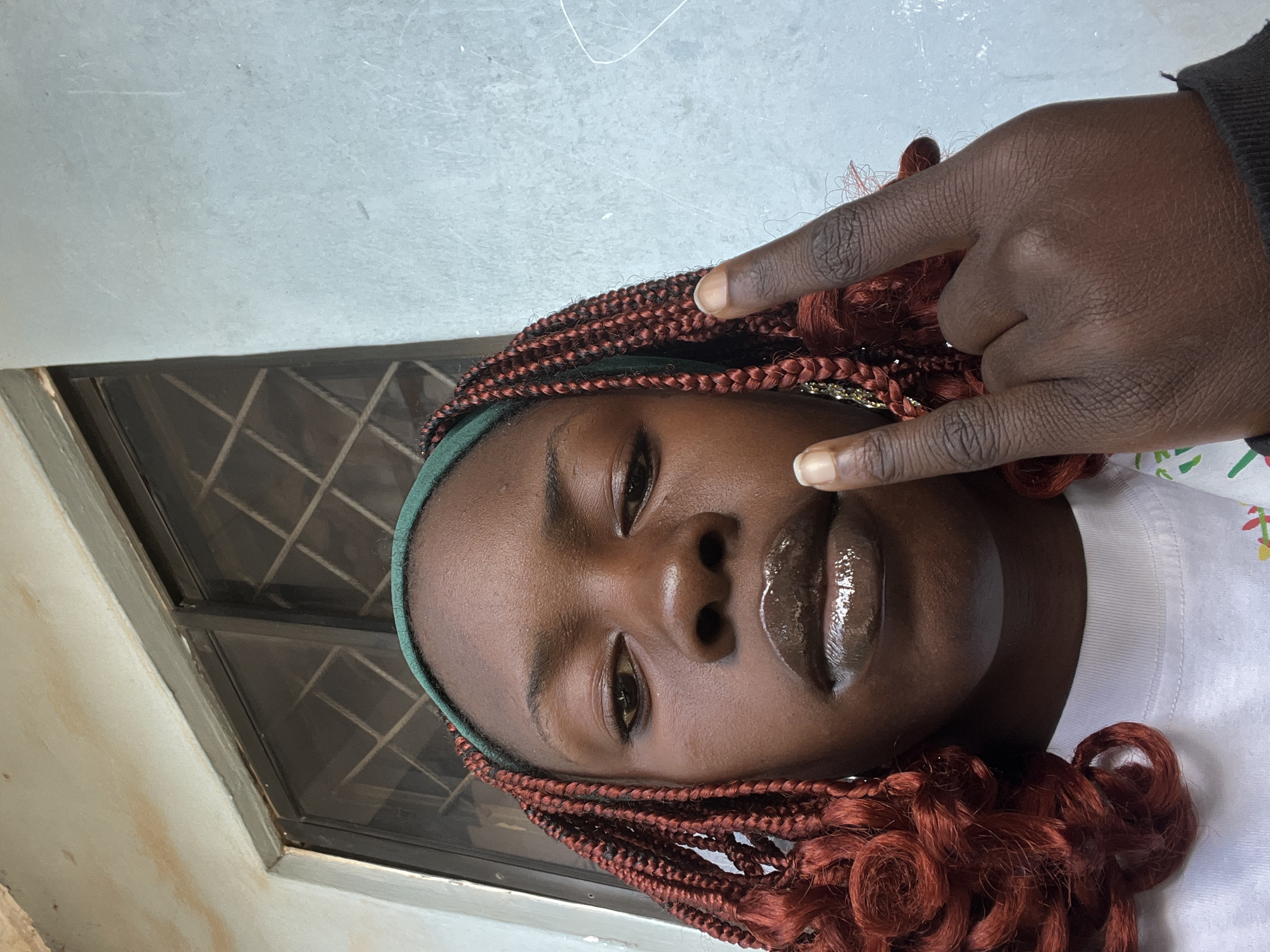 A close-up, selfie-style photograph shows a young Black person from the chest up, against a textured, light blue wall. The person is looking directly at the camera with a neutral expression. They have medium-dark skin, braided hair with reddish-brown highlights, and are wearing a green headband and a white t-shirt. Their lips are heavily glossed with a dark brown shade. Their right hand is raised, with the index and middle fingers extended in a "rock on" gesture, and their thumb resting on their middle finger. A small earring is visible in their left ear. Behind them, part of a window with a criss-cross grill is visible, above a light-colored, weathered concrete surface. The setting appears to be outdoors in an urban environment, possibly residential or commercial, in Jos, Nigeria.