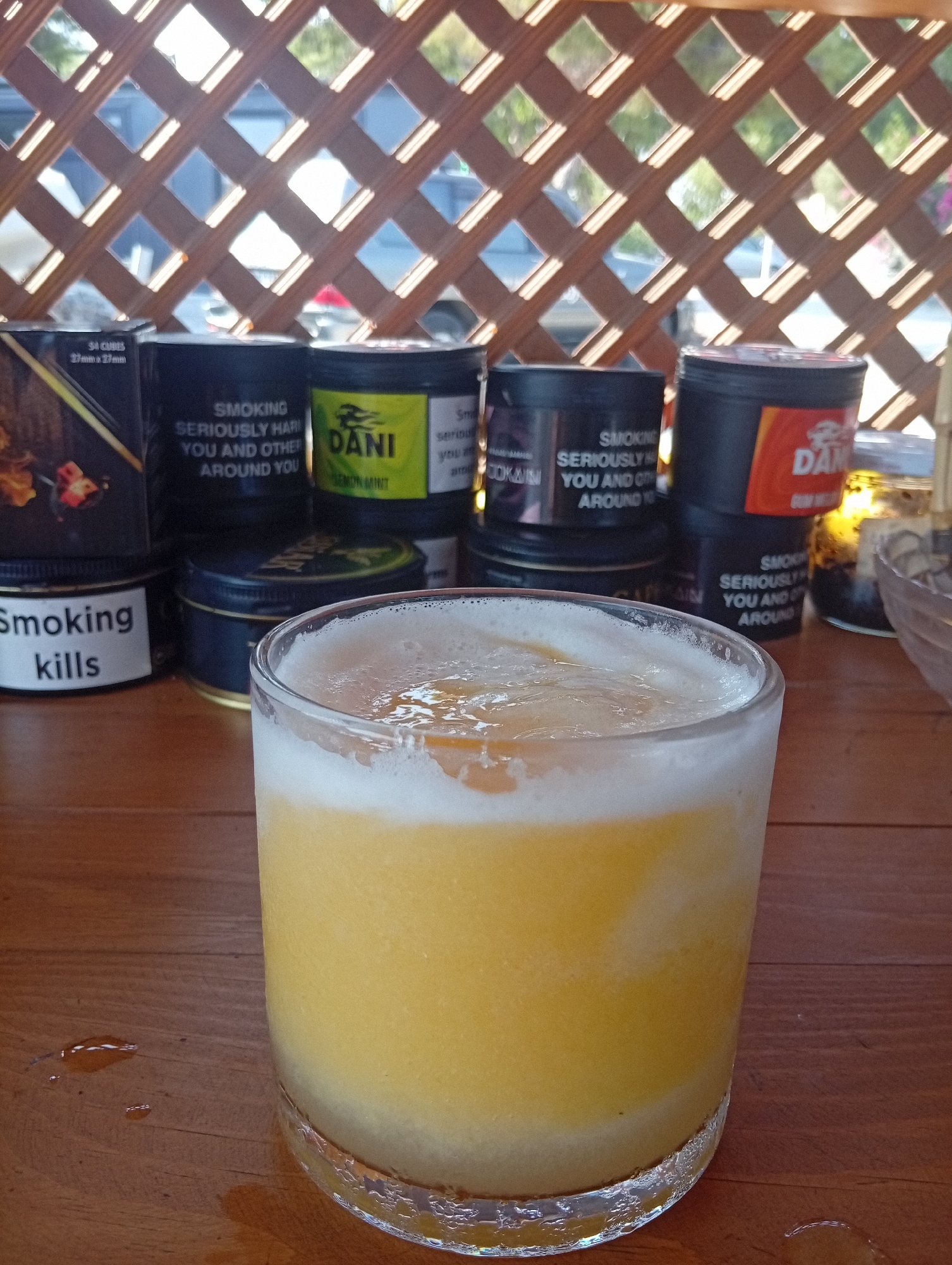 A close-up shot captures a frosted glass filled with a yellow beverage, likely a smoothie or cocktail, set against a wooden table. The drink features a foamy white layer on top, with a hint of orange garnish peeking through. Behind the glass, several cylindrical containers are visible, some with labels suggesting tobacco products. One label reads "Smoking Kills," and another displays the brand name "DANI" with "LEMON MINT" printed below. The background is a lattice structure with sunlight filtering through, casting diamond-shaped shadows. The setting appears to be outdoors, possibly a cafe or bar in Trikomo, Northern Cyprus, given the context of the request. The overall scene conveys a relaxed, possibly leisure-oriented atmosphere.