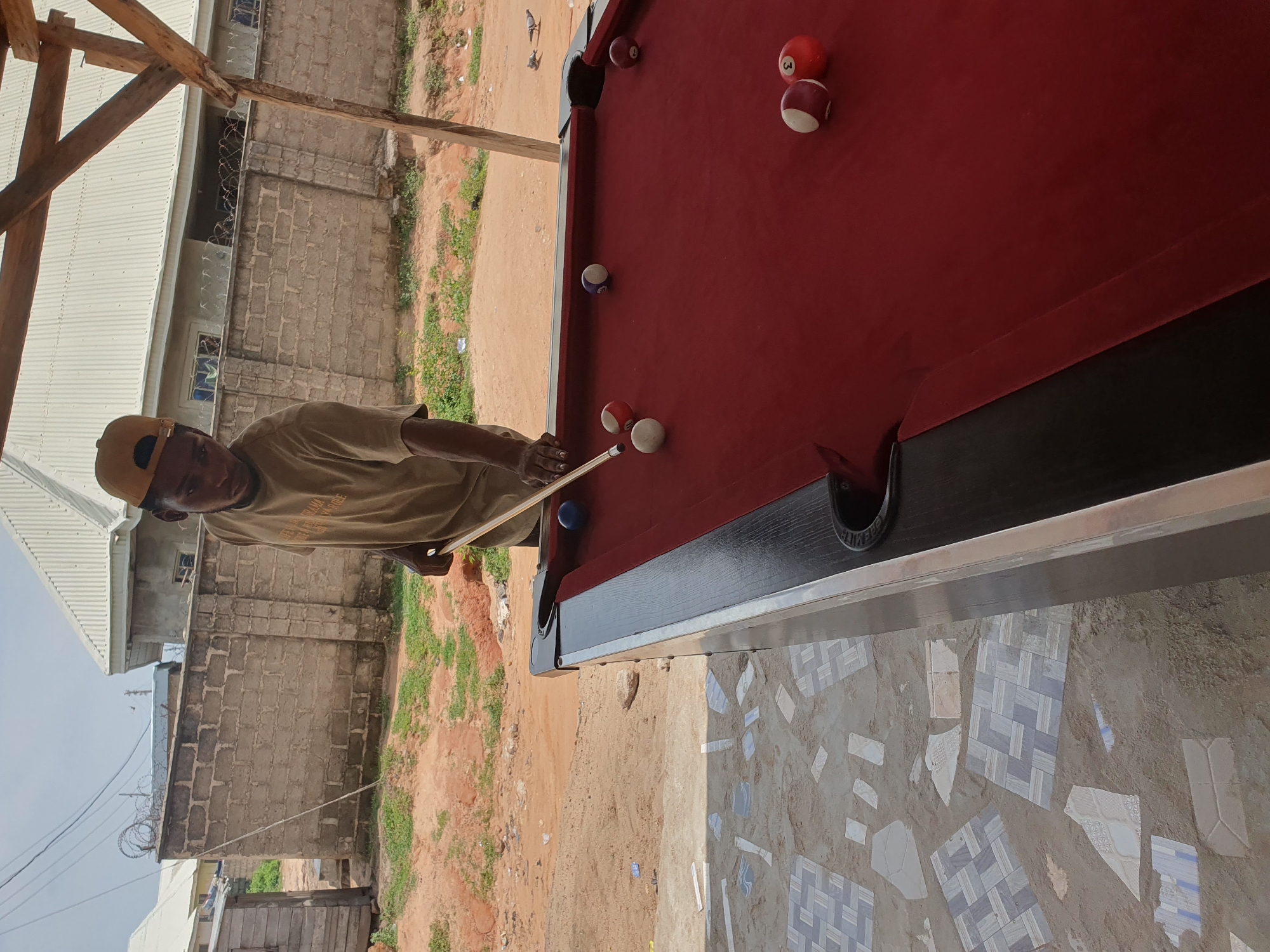 In Lokoja, Nigeria, a young man wearing a yellow cap and a tan t-shirt is playing billiards outdoors. He is positioned to strike a white billiard ball with a cue stick on a red felt-covered pool table. The table is surrounded by a black border and silver trim. Several other billiard balls are scattered on the table: a solid blue ball, a white ball with a red stripe, a ball with a purple stripe, two red and white striped balls, and a red ball with the number "3" on it. In the background, a low brick wall topped with barbed wire separates the billiard area from a dirt ground with scattered vegetation and debris. Beyond the wall, buildings with corrugated metal roofs are visible. The scene appears to be under a wooden structure, suggesting an open-air establishment. The ground in the foreground is a mosaic of broken tiles embedded in concrete.