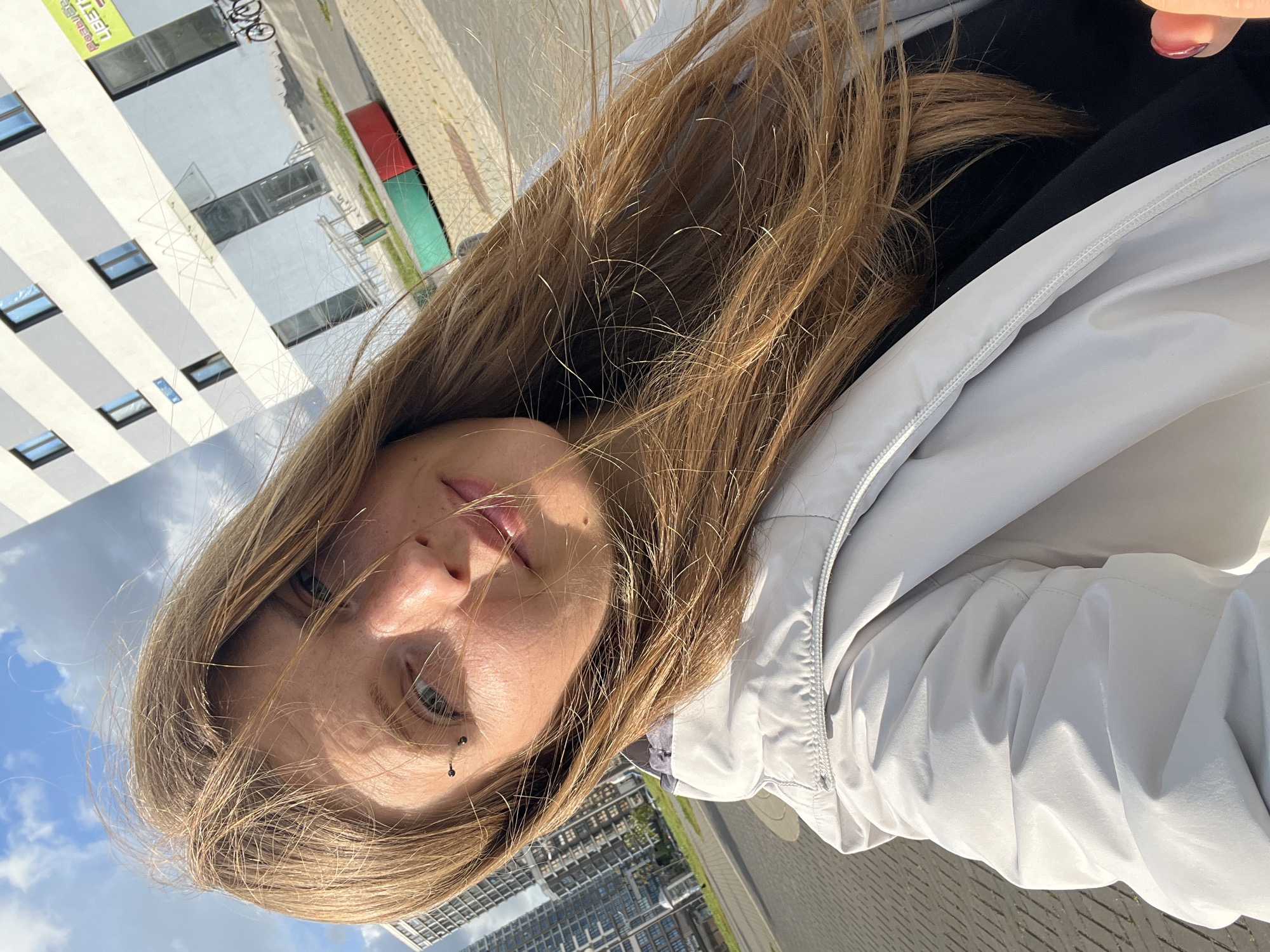 A close-up, high-angle selfie captures a young woman from the chest up, with her long, light brown hair blowing across her face. She is wearing a light grey hooded jacket over a black top. Her right eye is visible, along with her nose, lips, and cheek. She has a piercing above her right eyebrow. The background shows a modern urban environment with a white and grey building with blue-tinted windows, a paved walkway, and a street with vehicles. Tall residential buildings can be seen in the distance to the left. The sky is blue with scattered white clouds. The scene appears to be set in Minsk, Belarus, likely on a sunny day.