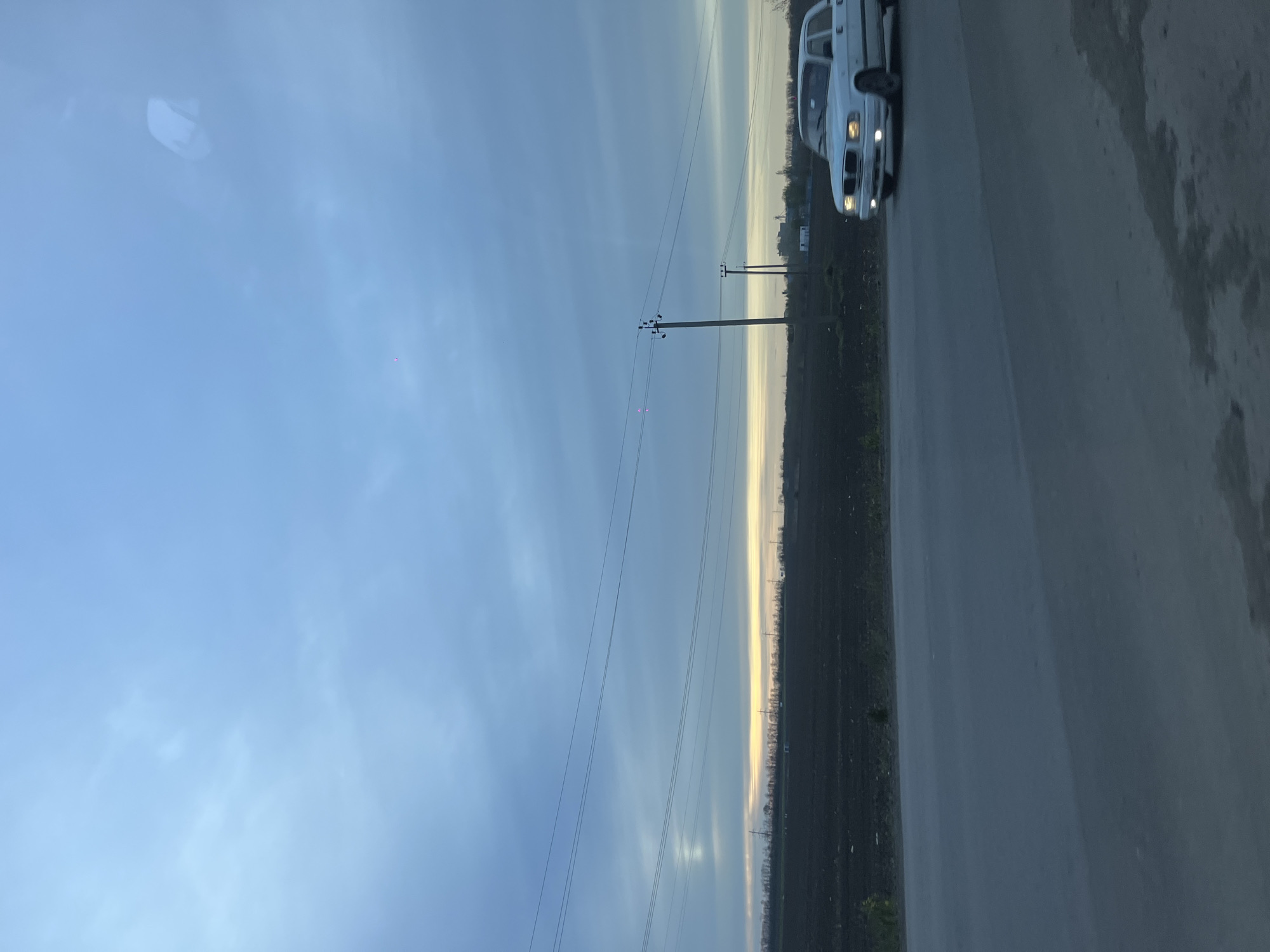 The image, captured from within a vehicle and rotated to a horizontal orientation, presents a twilight landscape. A pale blue sky, dotted with wispy clouds, occupies the majority of the frame, gradually transitioning into a narrow band of yellowish light along the distant horizon. This distinct horizon line is very low, suggesting a wide, open vista.
On the right side of the image, a paved road or shoulder extends into the distance. A white car, possibly a sedan, is partially visible on this road, its front end and illuminated headlights facing towards the viewer. To the left of the road, fields or cultivated land with discernible rows of vegetation stretch towards the horizon.
Several dark utility poles, connected by multiple power lines, are prominent in the mid-ground, receding into the distance. In the upper left portion of the sky, faint reflections or glares, likely from the interior of the capturing vehicle, are visible. A few small, bright pink points of light appear in the sky, possibly lens flares or artifacts.
The lighting conditions, characterized by the pale sky and the active car headlights, strongly suggest the time of day is either sunrise or sunset. The scene depicts a rural or semi-rural environment, consistent with the location context of Starominskaya, Russia. There is no visible text in the image.