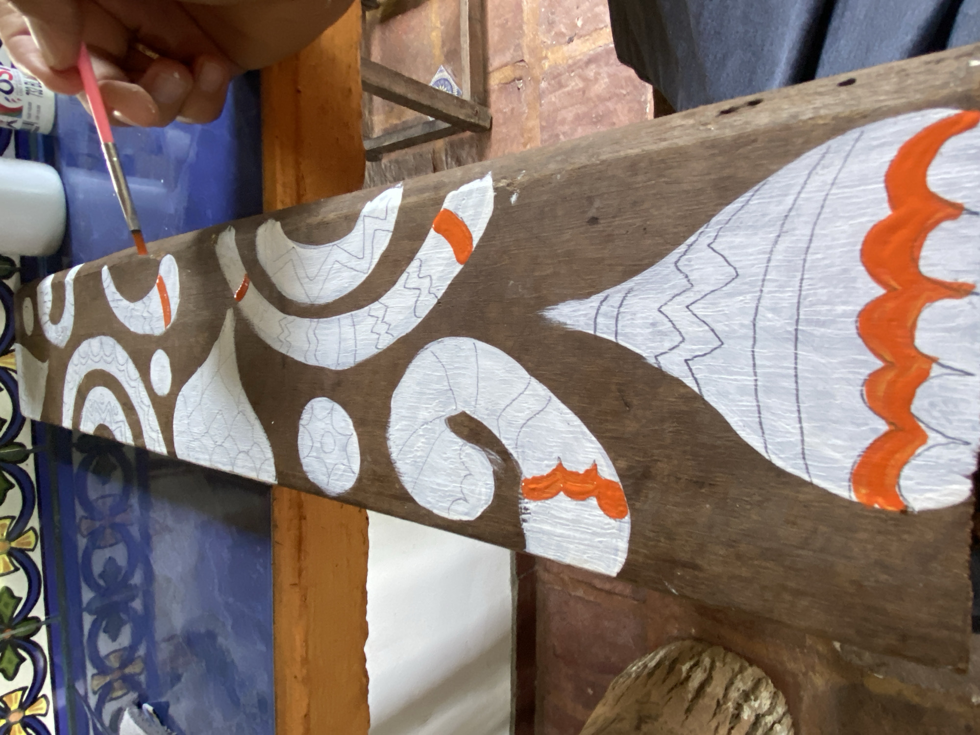An overhead shot shows a person's hands diligently painting a decorative pattern onto a long, horizontal wooden beam. The subject is holding a thin, pink-handled paintbrush and applying orange paint to a section of the intricate design.
The wooden beam, with its visible grain and rich brown color, serves as the canvas. A complex, organic pattern is partially painted on it in white, with some sections already having orange accents. The white painted areas feature detailed etched lines, including zig-zags and parallel strokes, suggesting depth or texture in the design. The artist is adding orange details to these white forms.
In the background, to the left, a portion of a colorful tiled surface is visible, featuring ornate blue, green, and yellow floral or geometric motifs. Adjacent to the tiles is a bright blue surface. A white plastic container, possibly for paint or water, is partially seen on the left. On one container, a red number "3" is faintly visible. To the right, another piece of wooden structure with a rough, textured surface is present. The setting appears to be an indoor or covered workspace, possibly in Los Velásquez, Venezuela, given the location context, and the style of the art may reflect local cultural motifs. The activity captures a moment of artistic creation, focusing on the detail and precision involved in painting the wooden beam.