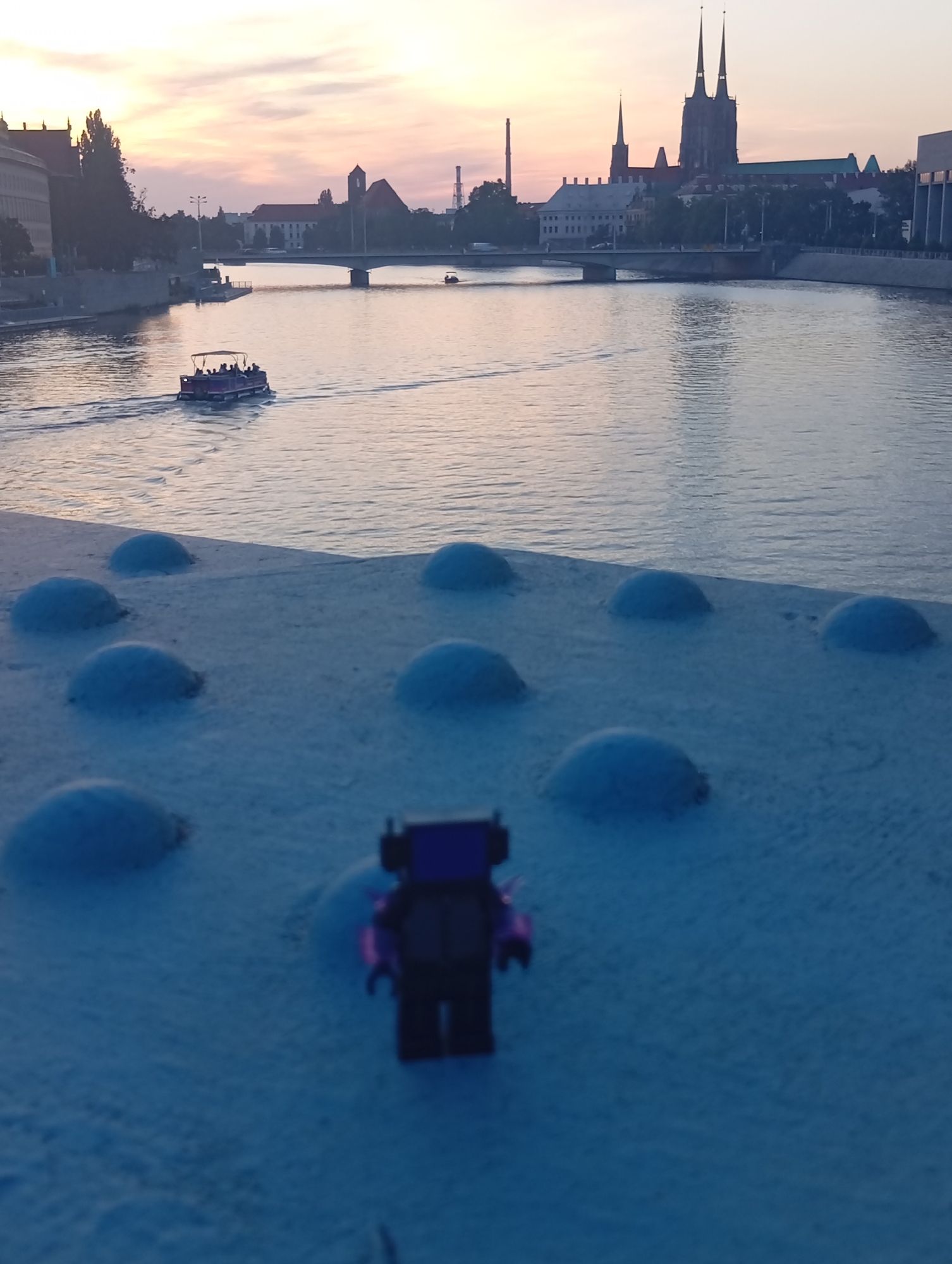 The image captures a waterfront scene at dusk, likely in Wrocław, Poland, given the prominent Gothic architecture resembling the Cathedral of St. John the Baptist in the distance. In the foreground, a white, textured surface is dotted with several rounded, blue mounds, possibly decorative elements on a bridge. A small, dark-colored Lego-style figurine with purple accents stands on this surface, facing away from the viewer. Across the calm, reflective water, a tour boat with a canopy and passengers is moving away from the viewer, leaving a wake. In the background, a cityscape with various buildings, bridges, and a tall industrial chimney is silhouetted against a soft, pastel-colored sky.