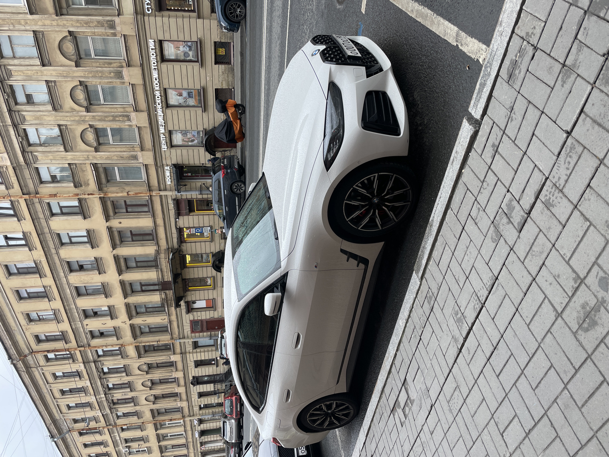The image shows a white BMW parked on a cobblestone street in Saint Petersburg, Russia. The building behind the car appears to be a multi-story structure with numerous windows and architectural details. A sign on the building reads "Центр Медицинской Косметологии", which translates to "Medical Cosmetology Center". Several other vehicles are visible in the background, including cars and what appears to be a covered motorcycle or scooter. The sky is overcast, and the car appears to be wet, suggesting it has recently rained.