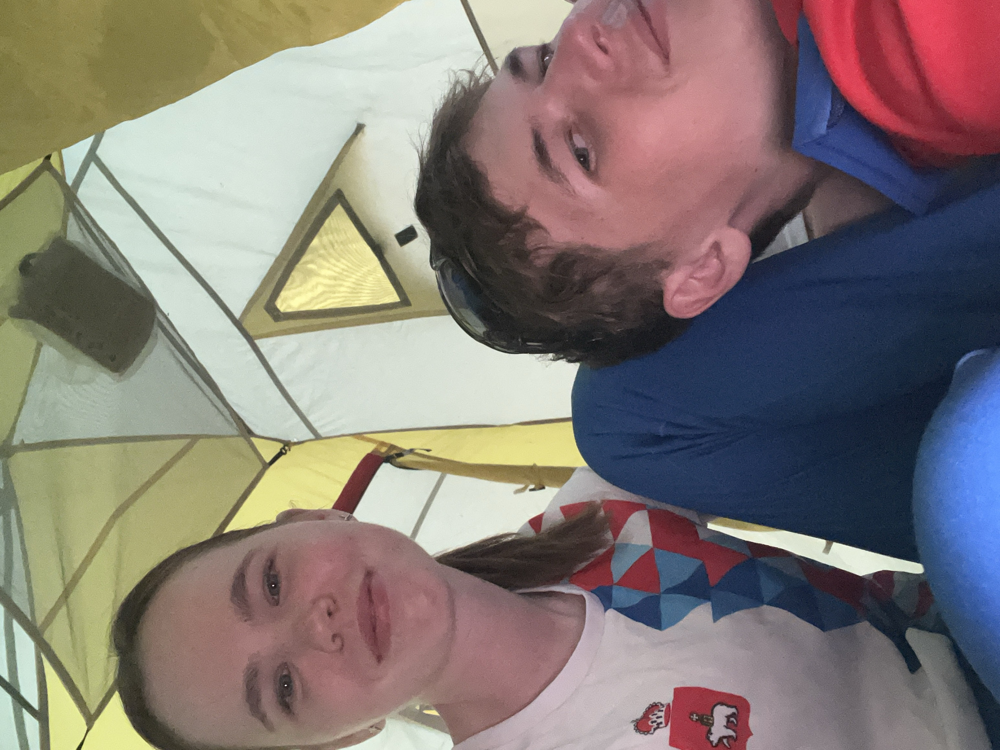 Two individuals are inside a tent. A woman with brown hair is looking at the camera. She is wearing a white t-shirt with a multicolored geometric pattern and a red patch with a white lion on it. A man with short brown hair and sunglasses is lying behind her. He is wearing a blue jacket and a red shirt. The interior of the tent is yellow and white. The location is Khvalynsk, Russia.