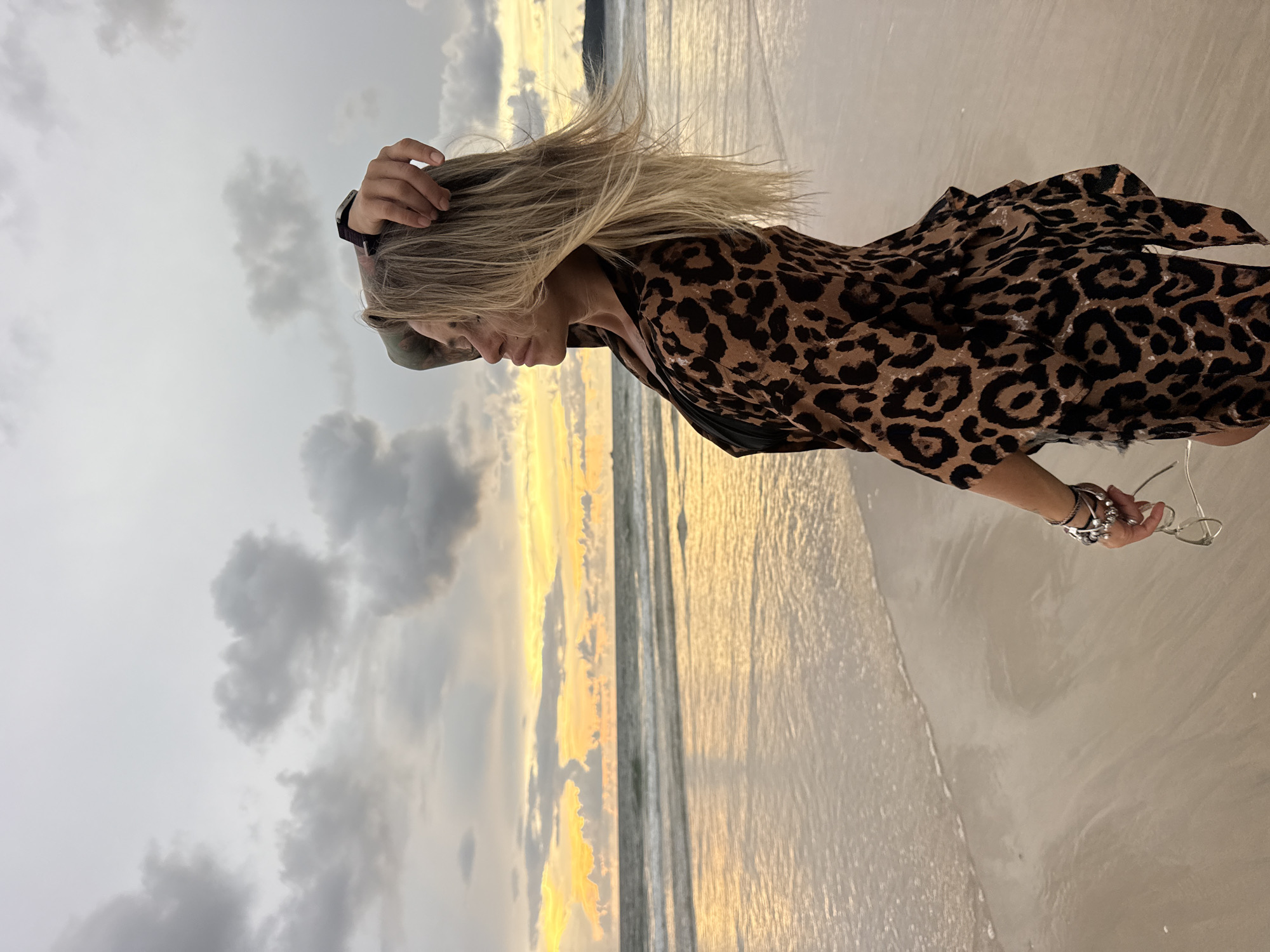 A woman stands on a sandy beach with the ocean in the background. She has blonde hair, which is being blown by the wind, and is wearing a leopard-print dress. Her left hand is touching her hair, and her right hand is holding a pair of glasses. She has tattoos on her left arm. The sun is setting, casting a warm glow over the water and the sky, which is filled with dramatic clouds. The waves are gently lapping at the shore. The overall mood of the image is serene and peaceful. The location is identified as Choeng Thale, Thailand.