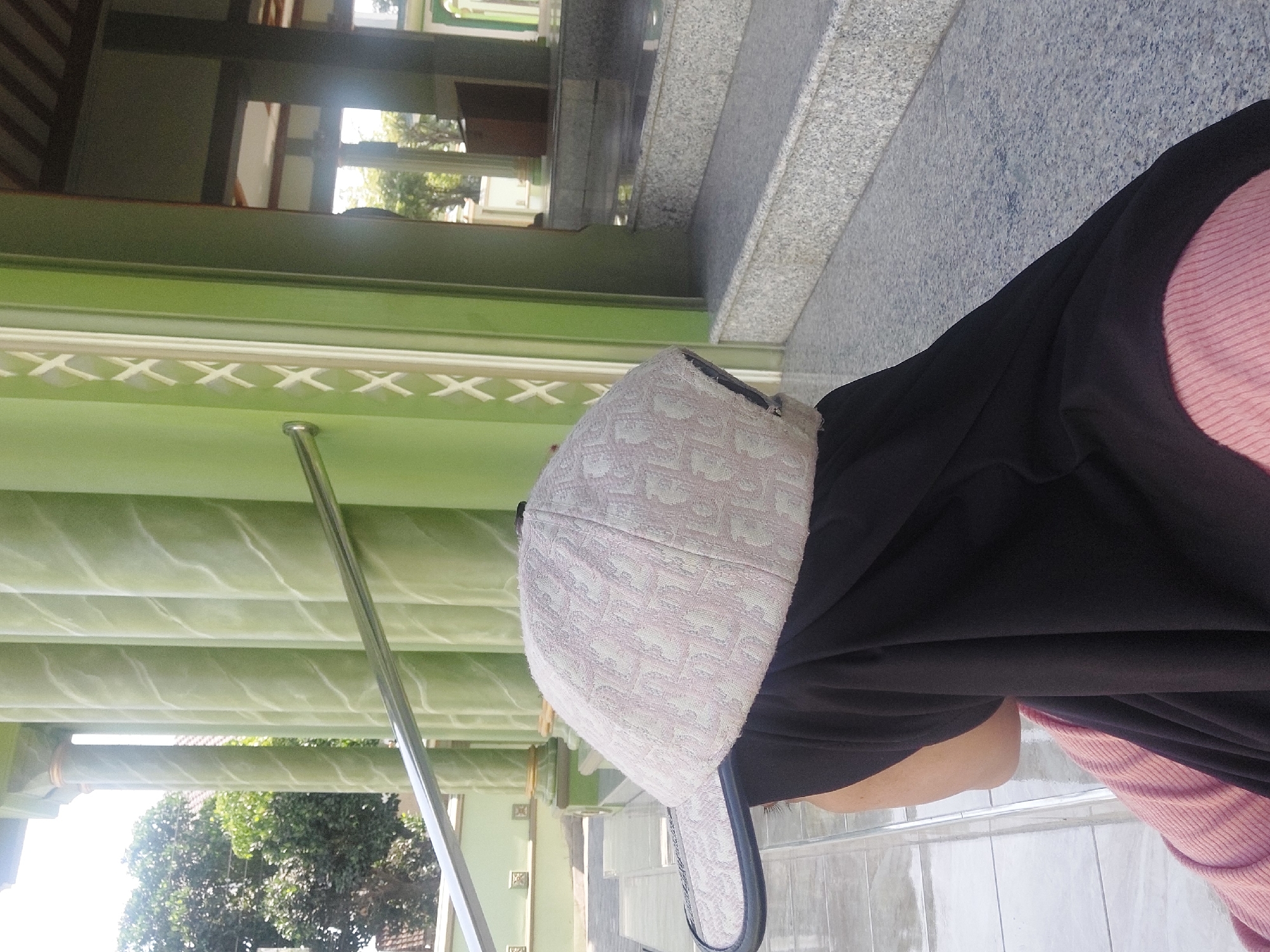 In Sedan, Indonesia, a person wearing a black hijab and a light-colored baseball cap with a repeating pattern is seated outside a mosque. The mosque has pale green pillars with white swirling decorative elements. In the background, there are steps leading up to a building with a wooden ceiling. Trees are visible in the distance.
