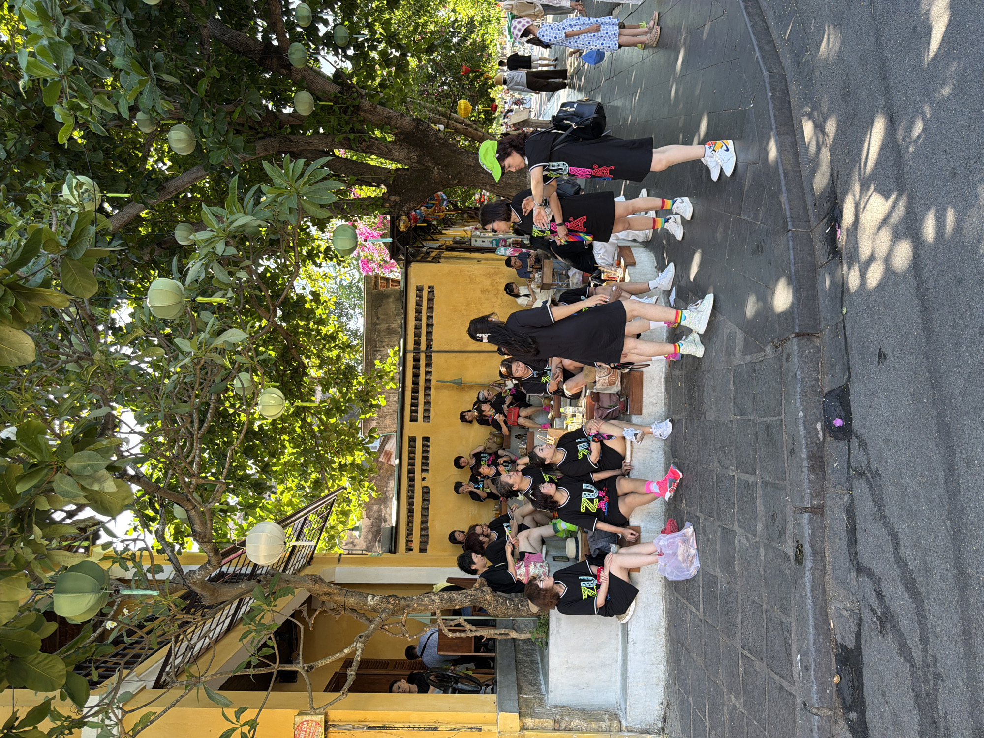 The image depicts a sunny outdoor scene in Hội An Ward, Vietnam, likely a pedestrian area with a yellow building and a tree-lined street. A group of women, many wearing matching black t-shirts with a "ZUU" logo, are gathered on a stepped walkway in front of the yellow building. Some are seated, while others are standing and interacting. The tree above them is decorated with several hanging paper lanterns in green and white. The ground is paved with dark stone slabs, and the street in the background shows more people walking and a few bicycles. The overall atmosphere suggests a casual gathering, possibly a break during an event or tour.