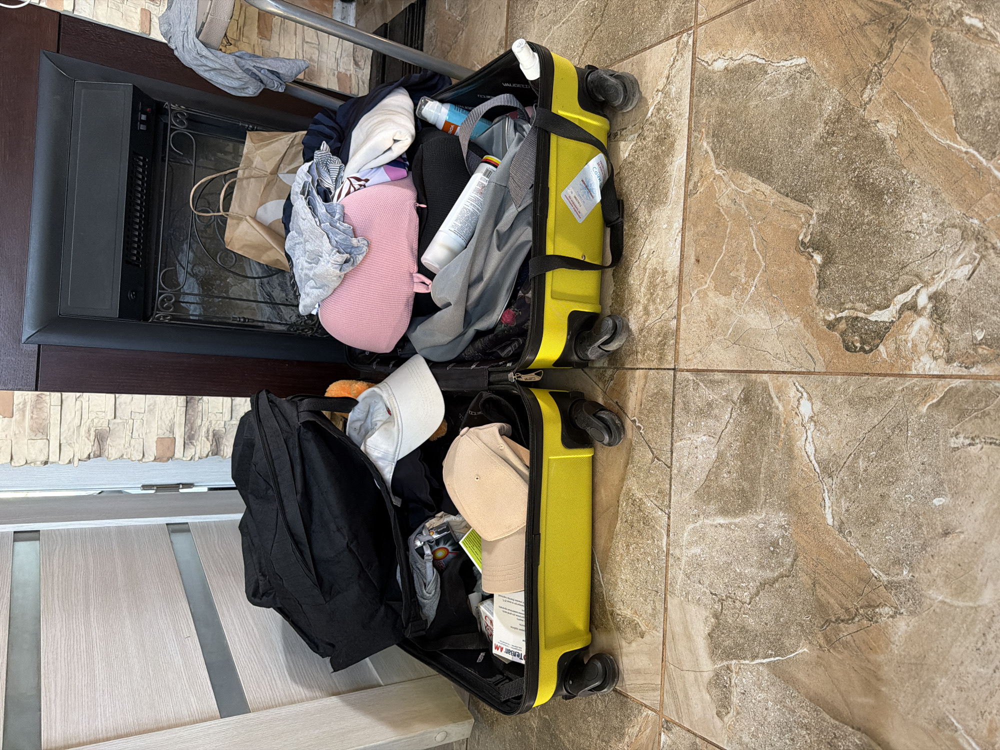 Two open yellow suitcases are on a tiled floor. The left suitcase is packed with clothing, including a black bag and a white cap. The right suitcase is also filled with clothing and toiletries, including a pink pillow and several spray bottles. In the background, a fireplace with a decorative grate is visible, along with a stacked stone wall and a wooden door. The scene appears to be indoors, likely in a residential setting. There are no people visible in the image. The location is identified as Shumki, Russia.