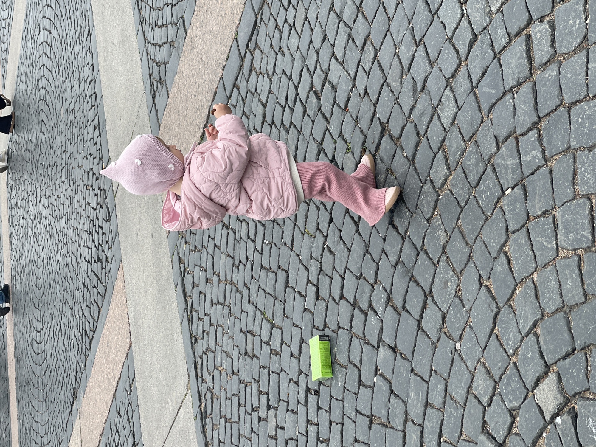 A young child dressed in a pink quilted jacket, pink pants, and a matching pink hat is lying on a cobblestone pavement. The pavement is made of dark grey cobblestones in a repeating pattern, interspersed with lighter grey stone borders. A green box-shaped object lies on the cobblestones near the child's feet. Part of an adult’s legs and shoes is visible in the corner of the image. The location appears to be in Saint Petersburg, Russia.