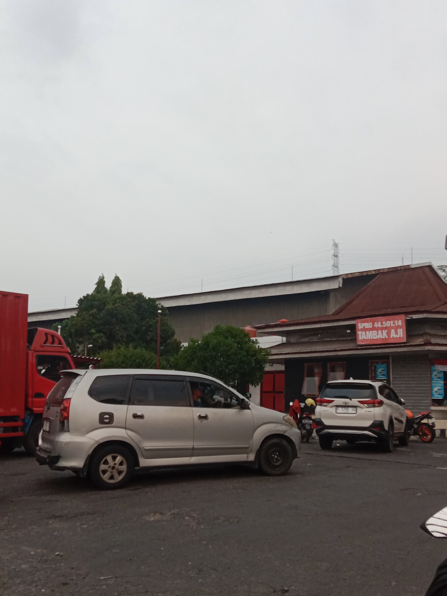 The image depicts a street scene in Semarang, Indonesia, with an overcast sky. In the foreground, a silver minivan is parked. To the left of the minivan, a red truck is partially visible. In the background, a building with a reddish-brown roof is prominent. A sign on this building reads "SPBU 44.501.14 TAMBAK AJI". Several other vehicles, including a white SUV and a motorcycle, are also present on the road. Trees and a utility pole are visible behind the building. The road surface appears to be asphalt.