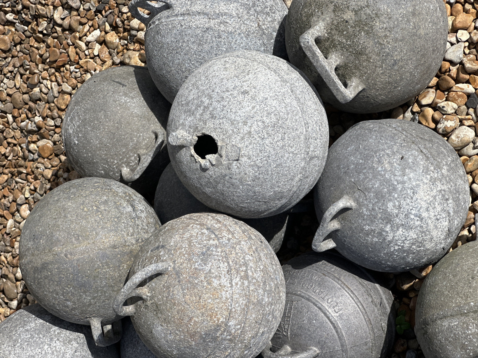 The image shows a close-up view of several grey, weathered, round objects, likely decorative spheres, resting on a bed of small, brown pebbles. Each sphere has a handle or loop attached to its surface. One sphere has a prominent hole. The scene's location is within the Folkestone and Hythe District of the United Kingdom, as indicated by the likely origin of such decorative objects for coastal locations. There are no people visible in the image. The objects are the central subject, and the environment consists solely of the pebbles. The interaction is simply the placement of the spheres on the ground.