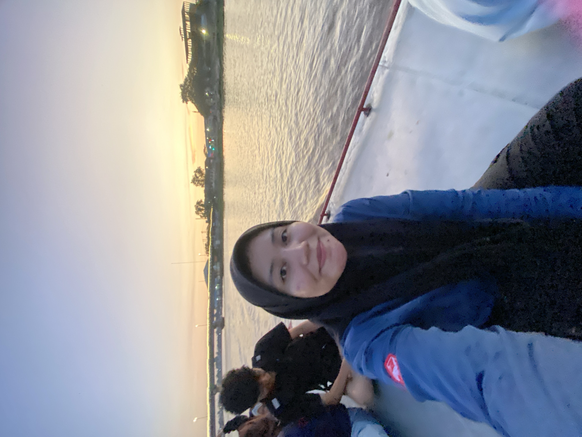 A medium shot shows a group of people on a boat at sunset in Banjarmasin, Indonesia. The main subject, a woman wearing a black hijab and a blue long-sleeved shirt, is in the foreground, smiling at the camera. Behind her, two men are visible, one wearing a black t-shirt and the other a dark shirt. The boat is on a wide river, with the orange glow of the setting sun reflecting on the water. In the background, a bridge with lights and buildings can be seen along the riverbank. The scene depicts a relaxed outing on the water during dusk.