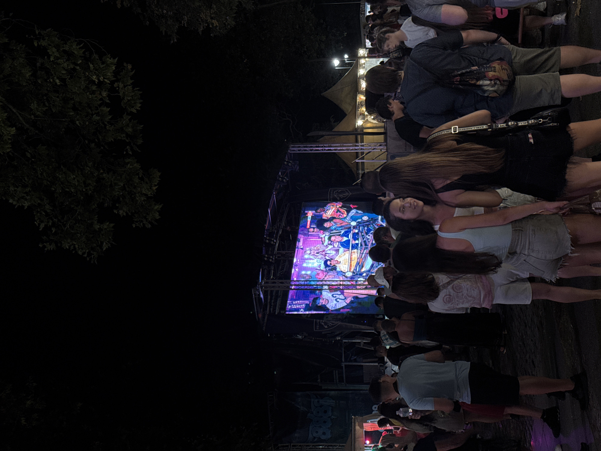 The image depicts an outdoor event at night in Burgas, Bulgaria. A large LED screen is set up on a stage, displaying a group of people. In front of the stage, a crowd of diverse individuals is gathered, some standing and talking, others watching the screen. The environment appears to be an open area, possibly a park or public square, with tents and trees visible in the background. The lighting is dim, with the screen and some spotlights illuminating the scene.
