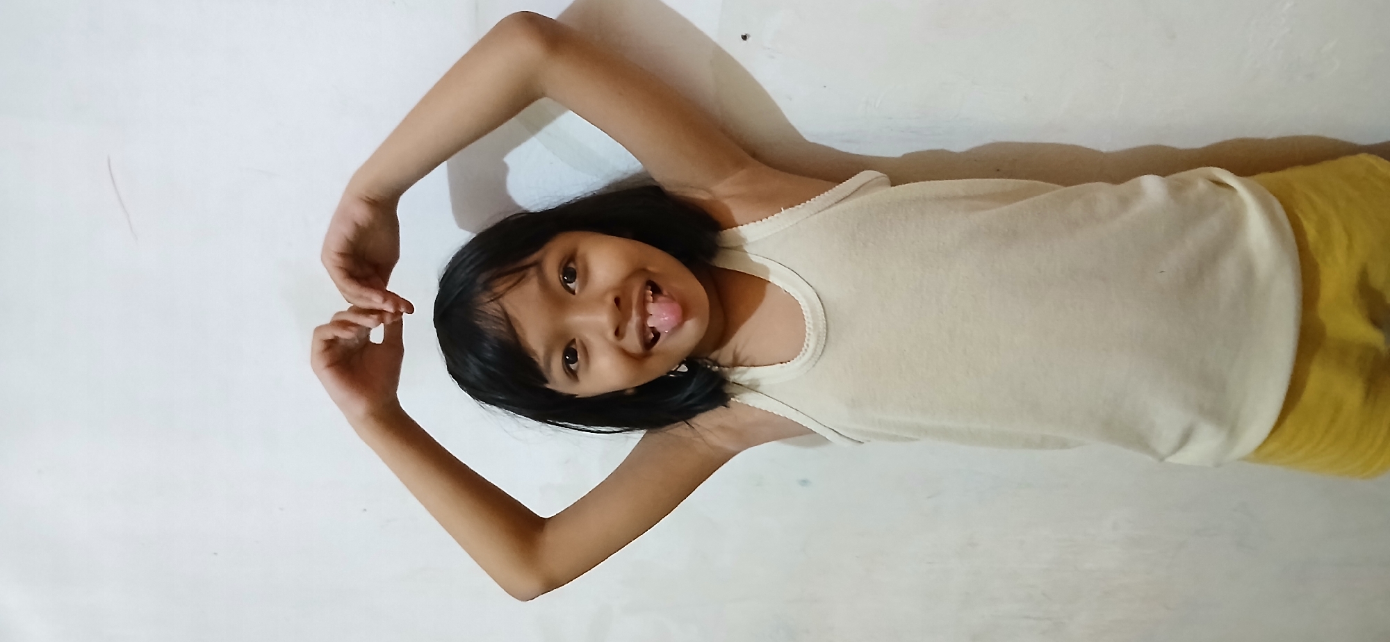 A young girl, likely of East Asian descent, stands against a plain, off-white wall, her arms raised and hands forming a heart shape above her head. She is wearing a light-colored sleeveless undershirt and yellow shorts. Her dark hair is cut in a bob, and she has dark eyes. Her tongue is sticking out, and she is smiling. The lighting suggests an indoor environment with a single light source casting subtle shadows. The wall appears to be textured, with some minor blemishes or marks. The composition is a head-and-shoulders shot, with the girl centered in the frame. The scene is simple and devoid of complex background elements, focusing entirely on the subject. There are no discernible objects or features that would help pinpoint a specific location within Samarinda, Indonesia.