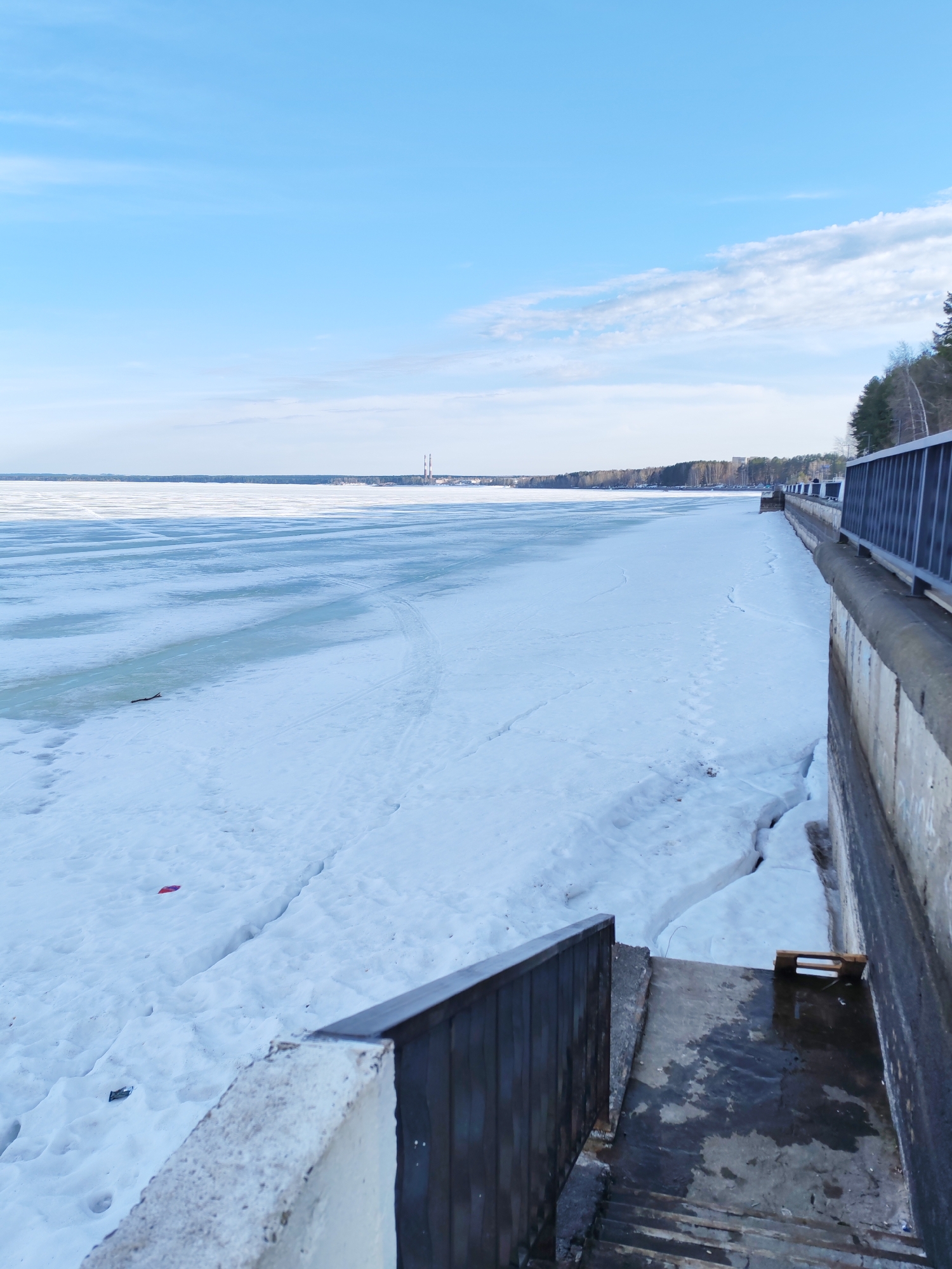 The image depicts a wintry scene in the city of Chaikovsky, Russia. A body of water, likely the Kama Reservoir, is covered with ice and snow. Tire tracks and footprints are visible on the snow-covered surface. In the distance, a forest borders the horizon, with what appears to be industrial smokestacks rising above the trees. To the right, a concrete embankment with a metal railing runs along the edge of the ice-covered area. A set of concrete stairs descends from the embankment towards the icy surface. The sky is a clear blue, with a few scattered clouds. The overall impression is a cold, overcast winter day.