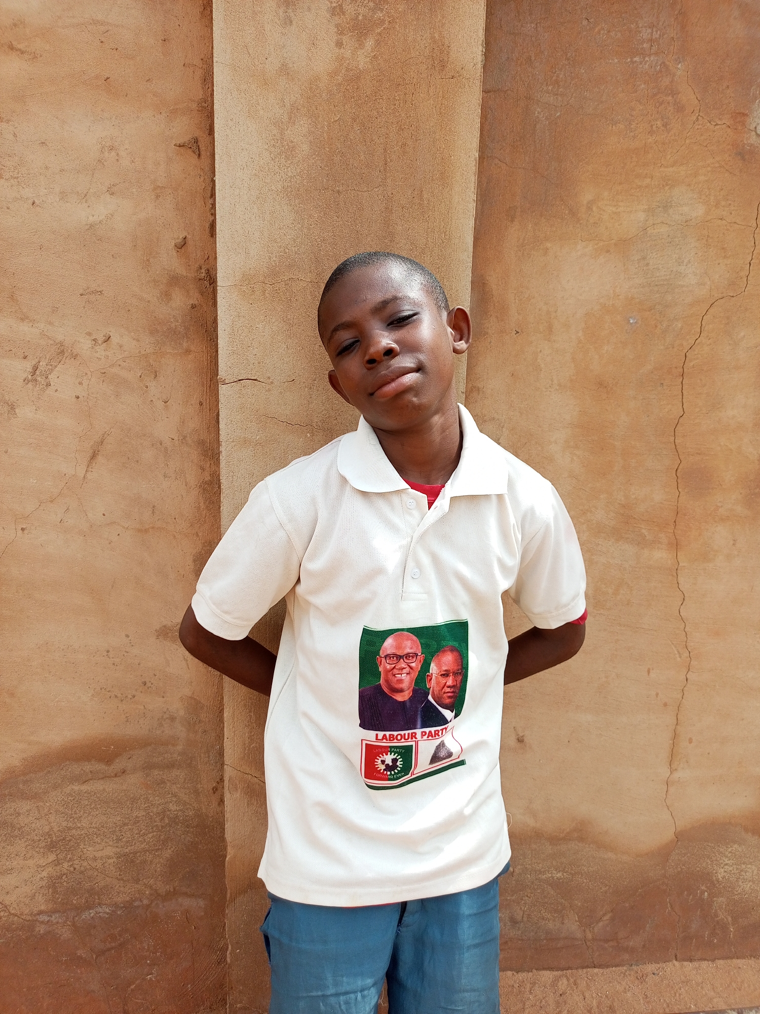 A young boy stands with his hands behind his back in front of a textured, tan-colored wall in Tempure, Nigeria. The boy wears a white polo shirt with a red collar and blue shorts. The shirt features a graphic of two men's faces, labeled "LABOUR PARTY." The wall is uneven and appears weathered, with some cracks and a vertical protrusion to the left of the boy. The lighting suggests it is daytime, with soft shadows indicating an overcast or diffused light source.