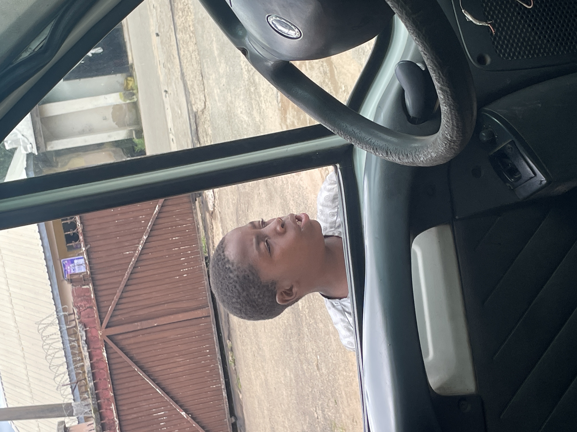 In Jos, Nigeria, a young boy looks out of the open window of a vehicle, his mouth slightly agape, suggesting he is speaking or reacting to something outside. He is wearing a grey shirt. The interior of the vehicle is visible, including a steering wheel and door panel. Outside the vehicle, a brown metal gate with diagonal bracing stands behind a low red wall topped with barbed wire. Beyond the gate, a building with a white roof and some signs is partially visible. The ground appears to be paved with concrete. The scene suggests an everyday moment on a street in Jos.