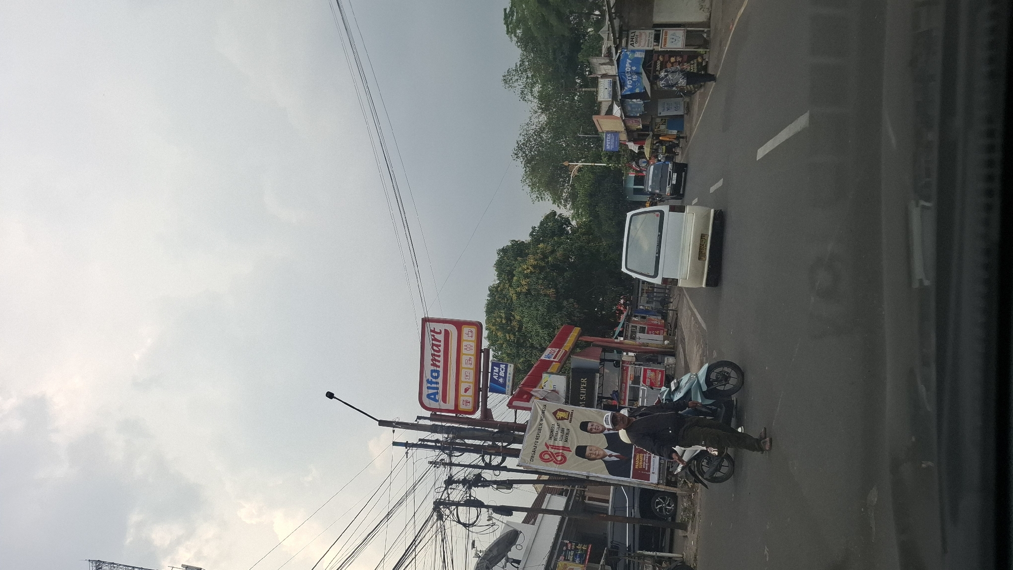 The image depicts a street scene in Sukasari, Indonesia, under an overcast sky. A prominent Alfamart convenience store sign is visible above a blue ATM BCA sign and other store signage. In the foreground, a man in a black jacket and green pants stands next to a teal-colored motorcycle on the left side of the road. He is facing towards the camera. Further down the street, a white minivan with a yellow license plate is driving away from the camera, followed by a dark-colored van. On the right side of the road, various small businesses are visible, including shops with blue awnings, one labeled "FOTOCOPY", and another with signs indicating "APOTER". Pedestrians can be seen near these businesses. The street is paved with asphalt and marked with white lane lines. Overhead power lines crisscross the sky.