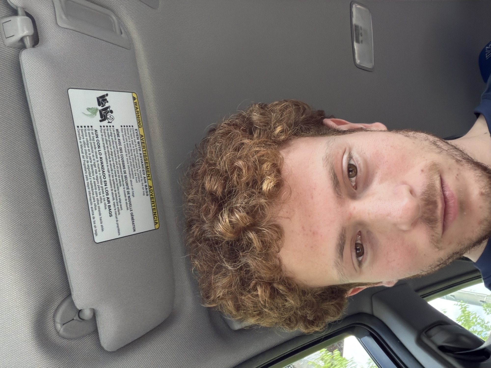 A selfie, rotated 90 degrees clockwise, shows a young man inside a car, looking directly at the camera with a neutral expression. He has light brown, curly hair and a short beard or stubble, and is wearing a dark blue shirt.
The setting is the car's interior, featuring a gray headliner and a prominent gray sun visor in the upper left. Attached to the sun visor is a multi-language warning label (English, French, Spanish) about advanced air bags. The text advises that children can be killed or seriously injured by air bags, states that the back seat is the safest place for children, prohibits rear-facing child seats in the front, and mandates the use of seat belts and child restraints. It also directs the reader to the owner's manual for more information.
A car ceiling light fixture is partially visible in the upper right. Through the car window in the lower right, green trees and a bright sky are visible, indicating it is daytime with good weather. The scene is likely set in Fort Lee, United States of America, consistent with the multi-language warning.