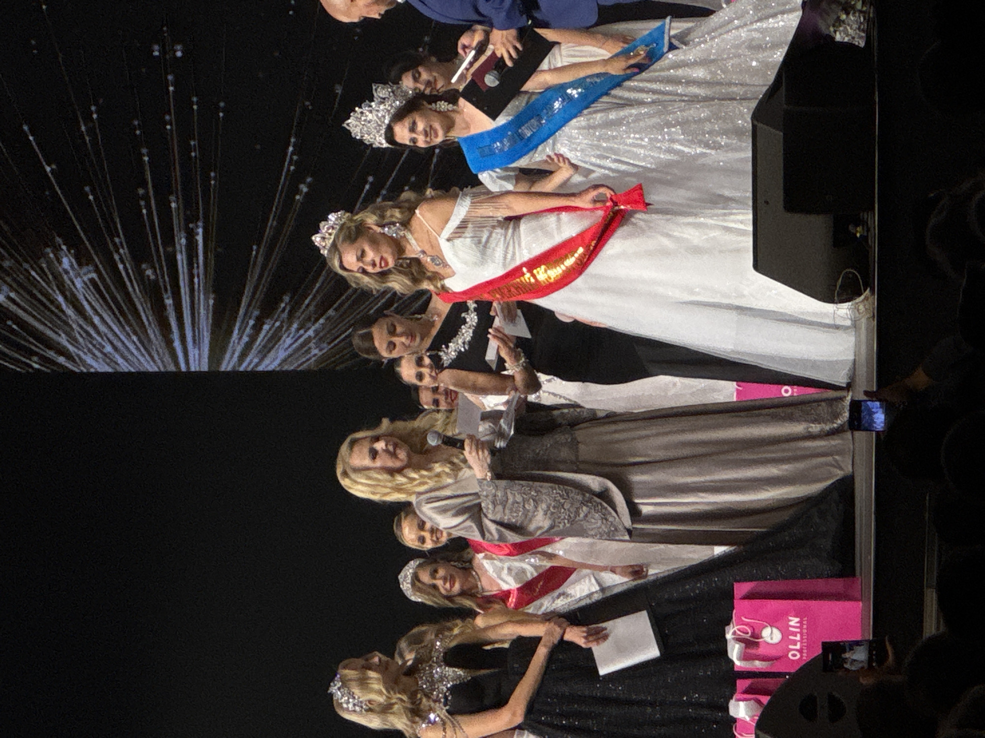 On a stage in an unspecified city, a group of women wearing crowns and long dresses are positioned in a line. The women are wearing red, white, and blue sashes with writing on them, and some are holding microphones and papers. A few men are present in the background, and the audience is visible on the right. A pink shopping bag is present in the lower right of the scene.