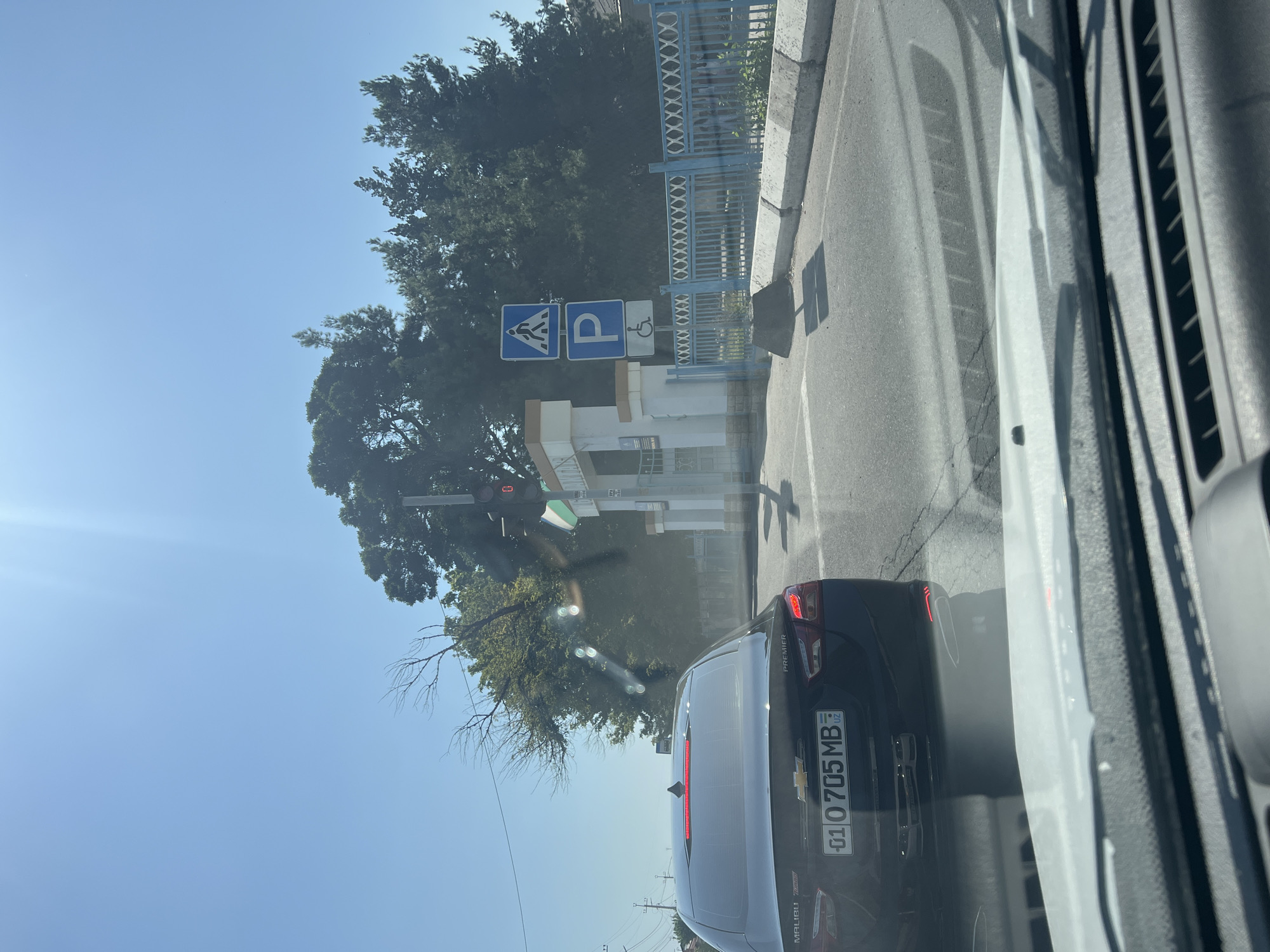 A dark sedan is driving down a street in Baytkurgan, Uzbekistan. The car has a license plate that reads "01 0705 MB UZ." In the background, there is a traffic light, a pedestrian crossing sign, a parking sign, and a sign for disabled parking. There are also trees and a blue fence. The sky is clear and blue.