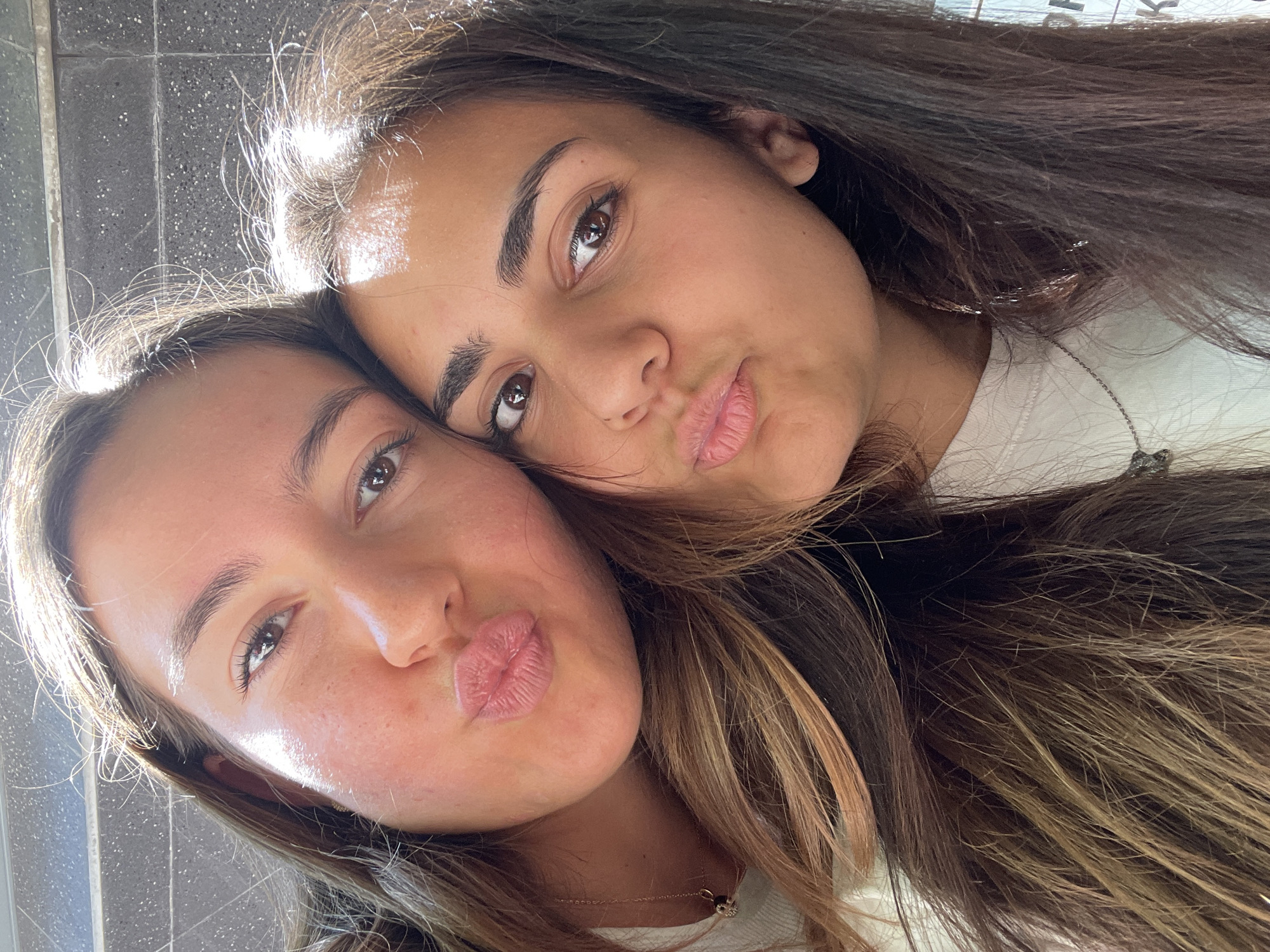 The image is a close-up selfie of two young women, likely friends, posing together. Both women have their lips pursed in a kiss-like gesture. The woman on the left has lighter skin and light brown hair. The woman on the right has darker skin and dark brown hair. They are both wearing casual white tops, and a delicate silver necklace is visible on the woman on the right. Their faces are illuminated by a bright light source, likely sunlight, which casts a warm glow. The background appears to be an outdoor setting with a textured surface, possibly a wall or pavement, with some vertical lines. There is no discernible information in the image to suggest a specific location within Burgas, Bulgaria.
