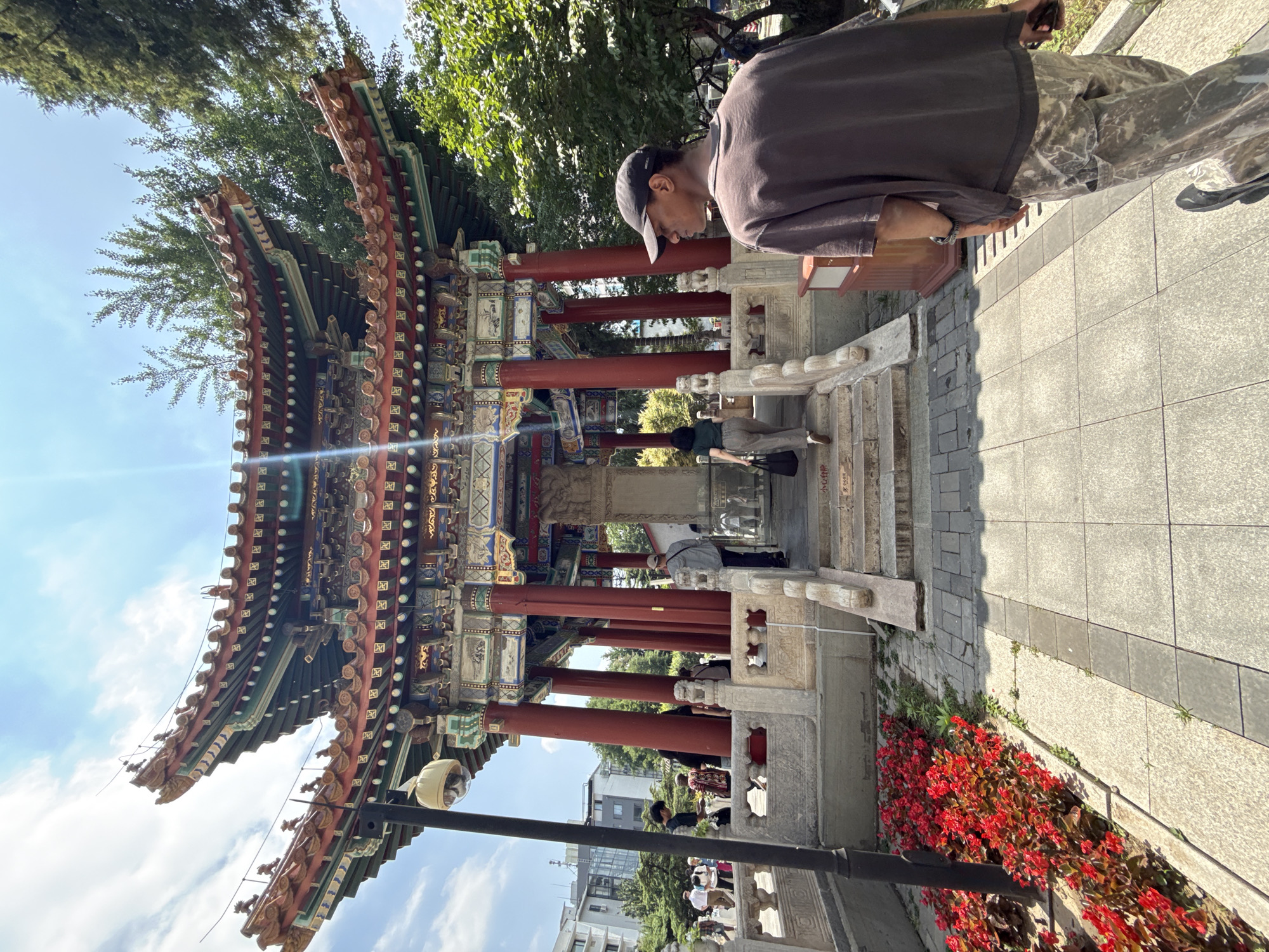The image depicts a traditional Chinese pavilion in Xicheng District, China, under a bright, sunny sky. The pavilion features multiple tiers of ornate, red and green tiled roofs with intricate decorative elements. Red-painted pillars support the structure, and a stone monument stands at its center. A balustrade with carved details flanks a set of stairs leading up to the pavilion.
In the foreground, a person wearing a brown t-shirt and camouflage pants walks away from the camera, looking to the right. To the left of the path, a vibrant bed of red flowers adds a splash of color to the scene. Several other people are visible in the background, walking towards and around the pavilion, suggesting a public space or tourist attraction. A security camera is mounted on a lamppost near the left side of the image. Buildings and trees form the backdrop, indicating an urban setting with green spaces. The overall scene suggests a visit to a historical or cultural site.