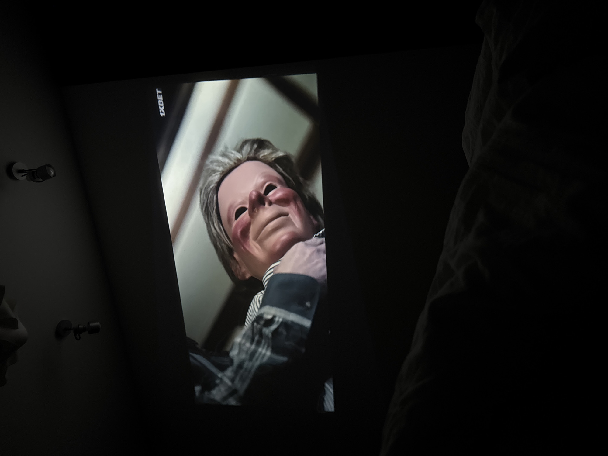 A screen displays an image of a human-like mask, perhaps plastic or latex, with blank eyes and a pinkish face. The mask is positioned at an angle, gazing down. It is wearing a checkered shirt and a white and black striped tie, with a black coat or suit jacket. Above the screen's image, to the left, are two small, dark cylindrical objects mounted on the wall, potentially lights or fixtures. The edge of a bed or sofa, draped with light-colored fabric, is visible in the dark background on the right side. The word "1XBET" is seen in the upper corner of the screen, and the image appears to be a still from a video. The location is Unknown, Unknown.