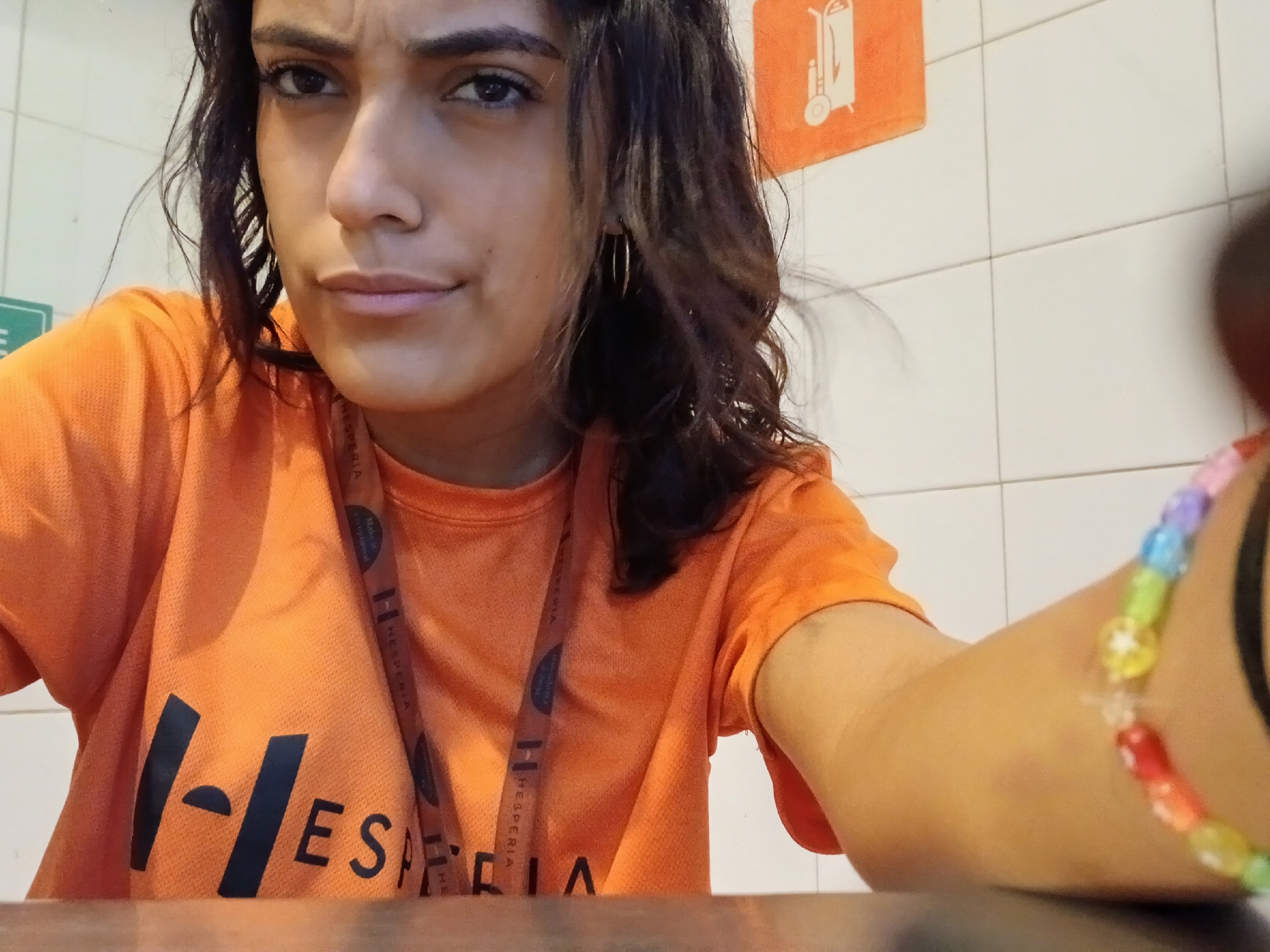 A close-up selfie shows a young woman, likely in her 20s or early 30s, looking directly at the camera with a serious or contemplative expression. Her dark, wavy hair frames her face, and she has dark eyes with slightly furrowed brows and neutral lips. She wears gold hoop earrings.
The woman is wearing an orange t-shirt featuring a prominent black "H-HESPERIA" logo on the front. An orange lanyard, also printed with "HESPERIA," hangs around her neck. Her right arm is extended towards the camera, revealing a colorful, multi-colored beaded bracelet and a black hair tie or band on her wrist.
The background consists of a white tiled wall, indicative of an indoor, possibly commercial or service, environment. An orange square sign with a white graphic of a hand truck or trolley is visible on the wall to her right. A small portion of a green sign is also visible on the left. Given the "Hesperia" branding on her uniform and lanyard, this image likely takes place within a Hesperia establishment in Playa El Agua, Venezuela. The indoor lighting does not suggest a specific time of day.