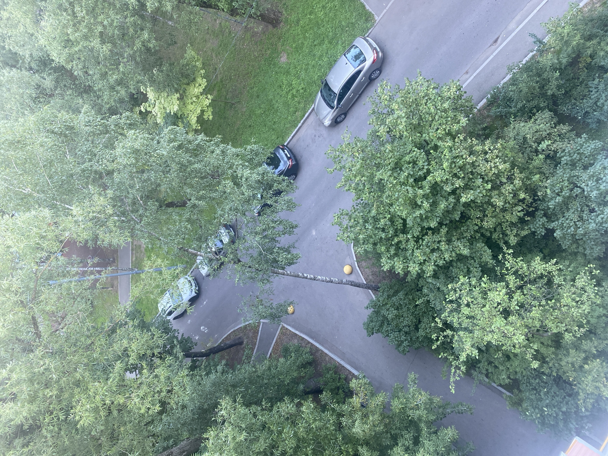 This is an aerial view of a residential courtyard in Moscow, Russia, featuring a paved road surrounded by lush green trees and grass. Several cars are parked on the road, including a light-colored SUV and a silver sedan. Yellow traffic bollards are visible on the pavement. The scene depicts a typical urban environment with abundant greenery.