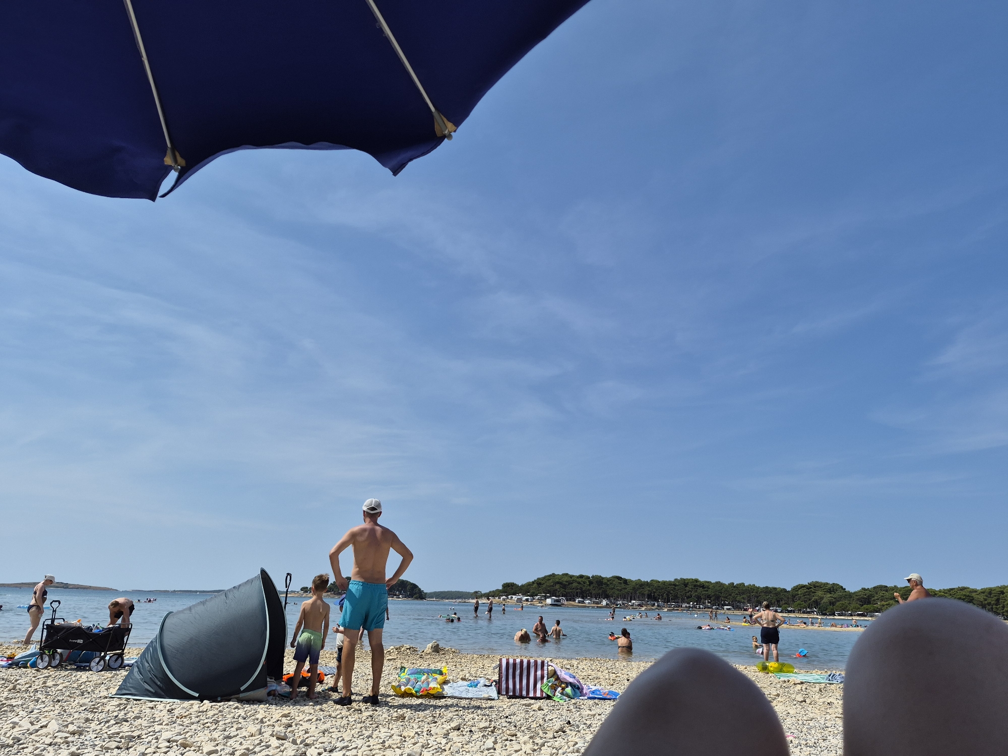 The image depicts a crowded beach scene in Općina Medulin, Croatia, on a sunny day. In the foreground, two pairs of legs are visible, suggesting the photographer is seated on the pebble beach. A dark blue beach umbrella partially obscures the sky. On the beach, a pop-up tent is set up, and a person stands near it, facing away from the camera. To the left, a woman wearing a swimsuit is seen next to a black wagon filled with beach gear. Several other people are present on the beach, some walking and others relaxing. The shallow, calm water is populated with numerous individuals swimming and wading. The background features a line of trees and vegetation, suggesting a coastal landscape with a developed area beyond the beach. The sky is clear blue with sparse white clouds.