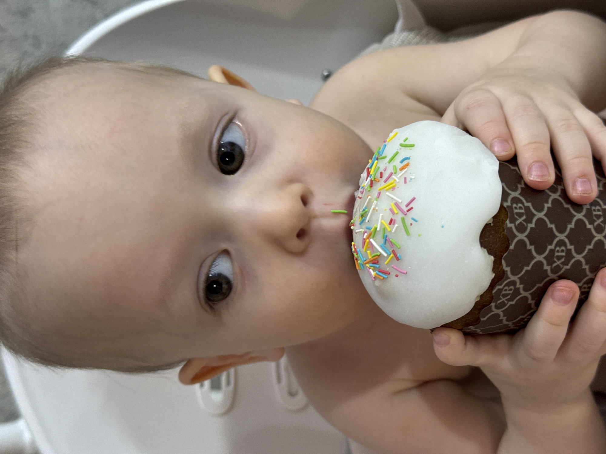 A close-up, angled, indoor shot features a baby with light skin and brown eyes, appearing to be around one year old, holding and tasting a decorated pastry. The baby's face, including the forehead, eyes, nose, and mouth, is clearly visible, with one small green sprinkle stuck to the lower lip. The baby's right hand is clutching the pastry, which is decorated with white icing and colorful sprinkles. The pastry itself has a patterned, dark brown base with the letters "XB" repeating across it. The background is blurry, revealing a white surface suggestive of a sink. The image likely originates from a domestic setting, but there is no information to confirm that it was taken in Chaikovsky, Russia.