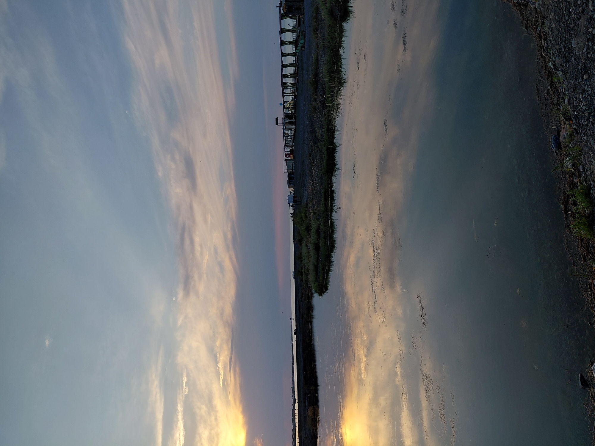 The image depicts a tranquil scene at sunset in Akshi, Kazakhstan. The sky is a canvas of soft hues, with wispy clouds painted in shades of pale pink, orange, and blue. A faint crescent moon is visible in the upper left portion of the sky.
Dominating the right side of the image is a calm body of water, reflecting the colors of the sunset and the sky like a mirror. The water's surface is smooth, with subtle ripples creating distorted reflections of the clouds. Along the edge of the water, a strip of land is visible, covered with pebbles and sparse green vegetation.
On the left side of the image, a low horizon line separates the sky and water. In the distance, a linear arrangement of structures is visible, possibly buildings or a pier, stretching along the shoreline. These structures are silhouetted against the twilight sky. A few boats might be present, but they are not clearly discernible.
The overall mood of the image is one of peace and serenity, characteristic of a sunset. The soft lighting and gentle reflections create a calming atmosphere. The time of day is clearly sunset, indicated by the warm, golden light illuminating the clouds and their reflection. The location is identified as Akshi, Kazakhstan. No text is visible in the image.