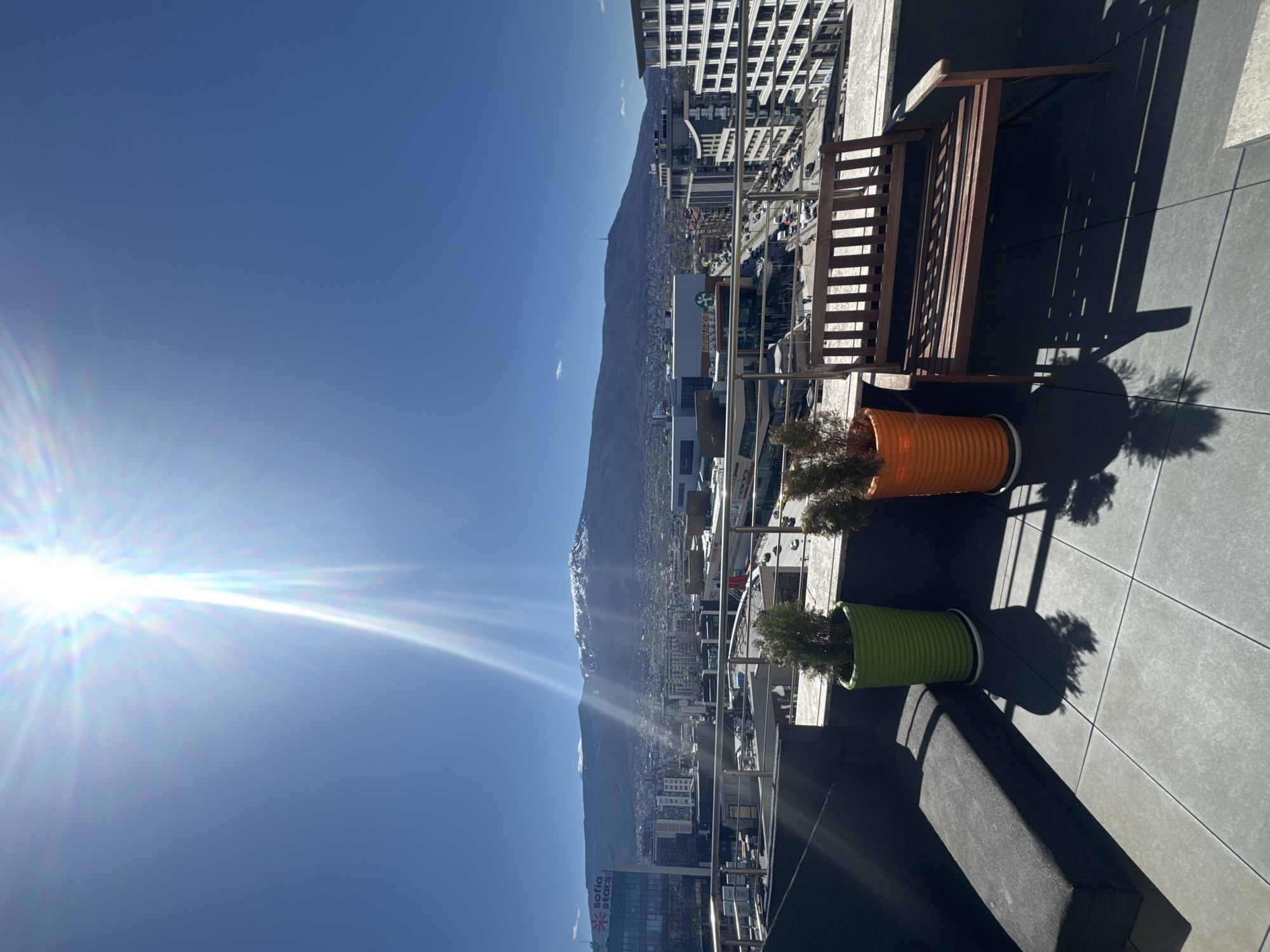 A long shot, taken from a high vantage point such as a rooftop or balcony, shows a scene of urban Sofia, Bulgaria, against the backdrop of a clear blue sky and a mountain range. The sun is shining brightly, causing a lens flare. In the foreground, there are two potted plants, one in orange and one in green, alongside a wooden bench. The cityscape includes various buildings, from apartments to commercial structures. At the bottom of the frame, the signage for "Sofia Stars" is visible, indicating the location within the city. The ground is tiled in a gray square pattern. The viewpoint offers an expansive view, showcasing both the urban environment and the natural landscape of Sofia.