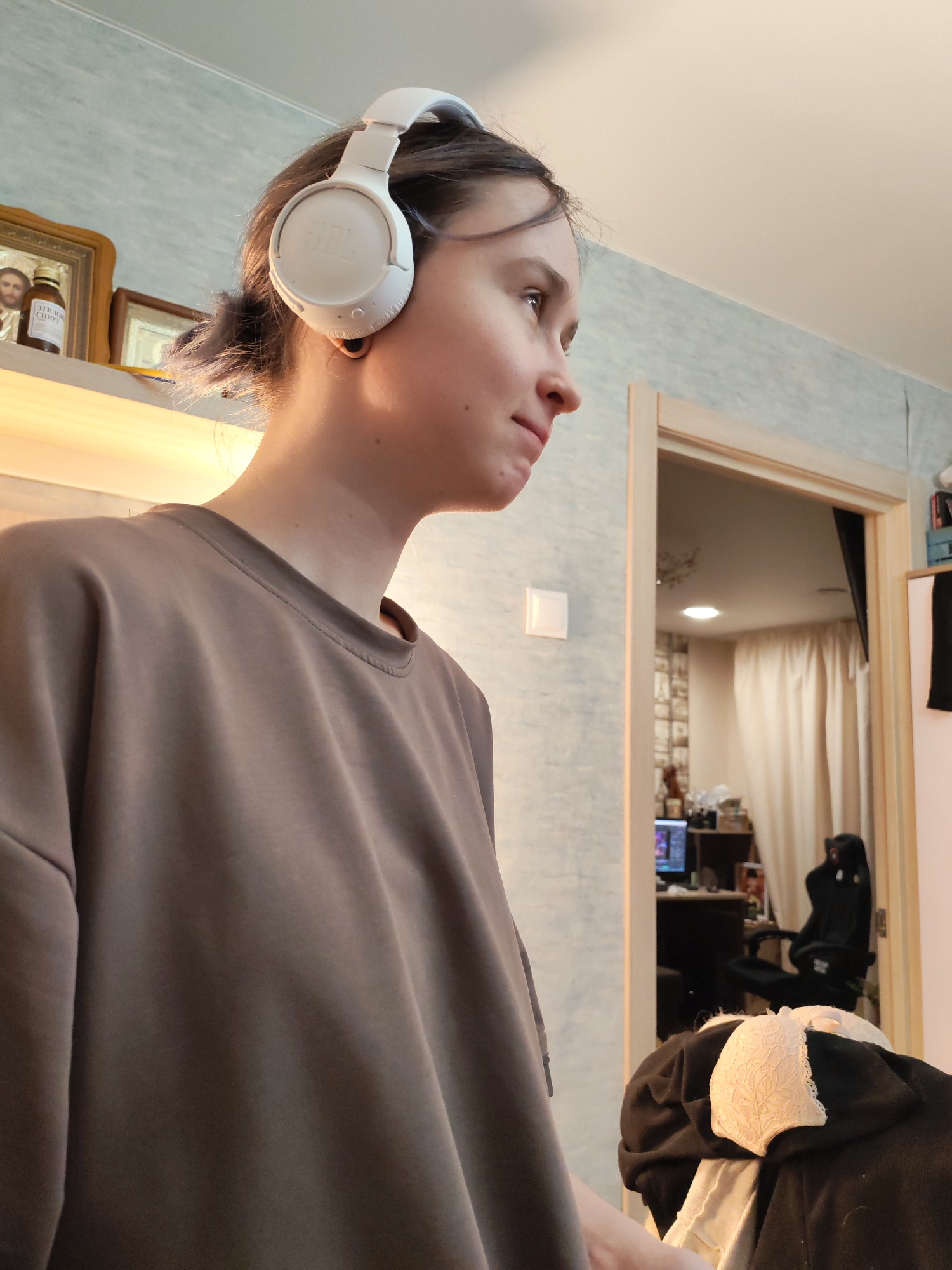 The image presents an indoor scene located in Chaikovsky, Russia. A young individual, likely female, is captured from the chest up in a right-profile view, wearing a solid-colored, loose-fitting taupe long-sleeve shirt and white JBL over-ear headphones. A gold-colored hoop earring is visible in her left earlobe. Her dark hair is pulled back, with some strands visible around her ear. She appears to be looking slightly upwards and to the right.
The immediate background features a light blue wall with a subtle textured pattern. Above the individual's left shoulder, on a light-colored shelf, a gold-framed religious icon depicting a bearded man is displayed alongside a brown bottle labeled in Cyrillic "МИРРОВИЙ СПИРТ" (Myrrh Spirit). A second, smaller framed item is partially visible.
To the right of the individual, a light-colored wooden doorframe opens into an adjacent room. A white square light switch is mounted on the wall to the left of this doorway. Through the doorway, parts of another room are visible, including light-colored curtains, a black gaming chair, and a desk setup with a computer monitor displaying illuminated graphics. Various other objects are arranged on the desk or shelves. A recessed ceiling light provides illumination in this background room. In the lower right foreground, a pile of clothing is visible, including a black garment and a white lace bra.