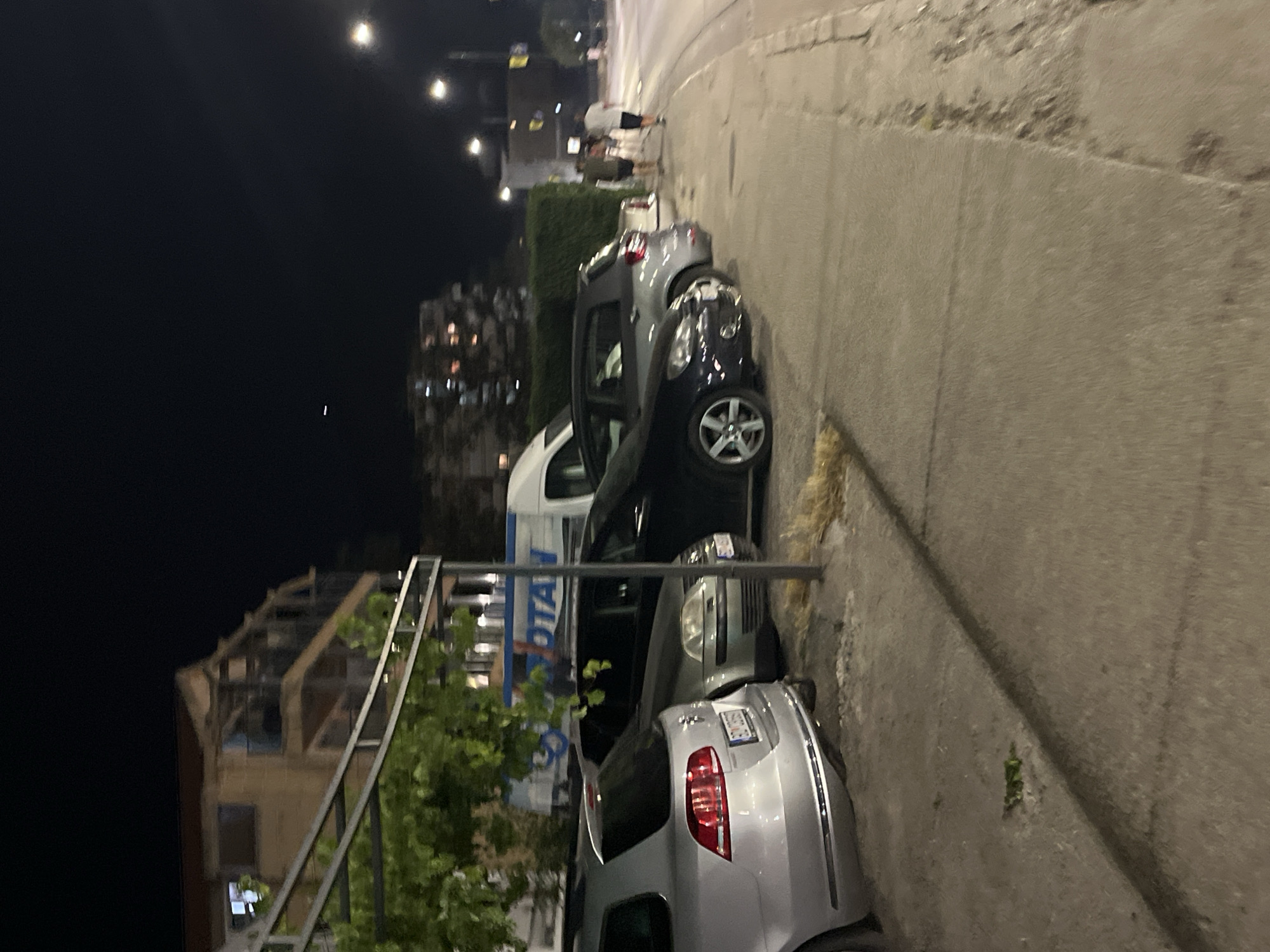 At night, in Budva, Montenegro, parked cars line a sidewalk next to a street. On the left, a silver station wagon is visible, followed by a dark sedan and a smaller gray car. Further down the sidewalk, two individuals, a man and a woman, are walking away from the camera. In the background, buildings and streetlights illuminate the scene. The environment suggests an urban setting with commercial activity, as indicated by a large truck with "OTAN" visible on its side, possibly a delivery vehicle. The overall atmosphere is calm and ordinary for a nighttime city environment.
