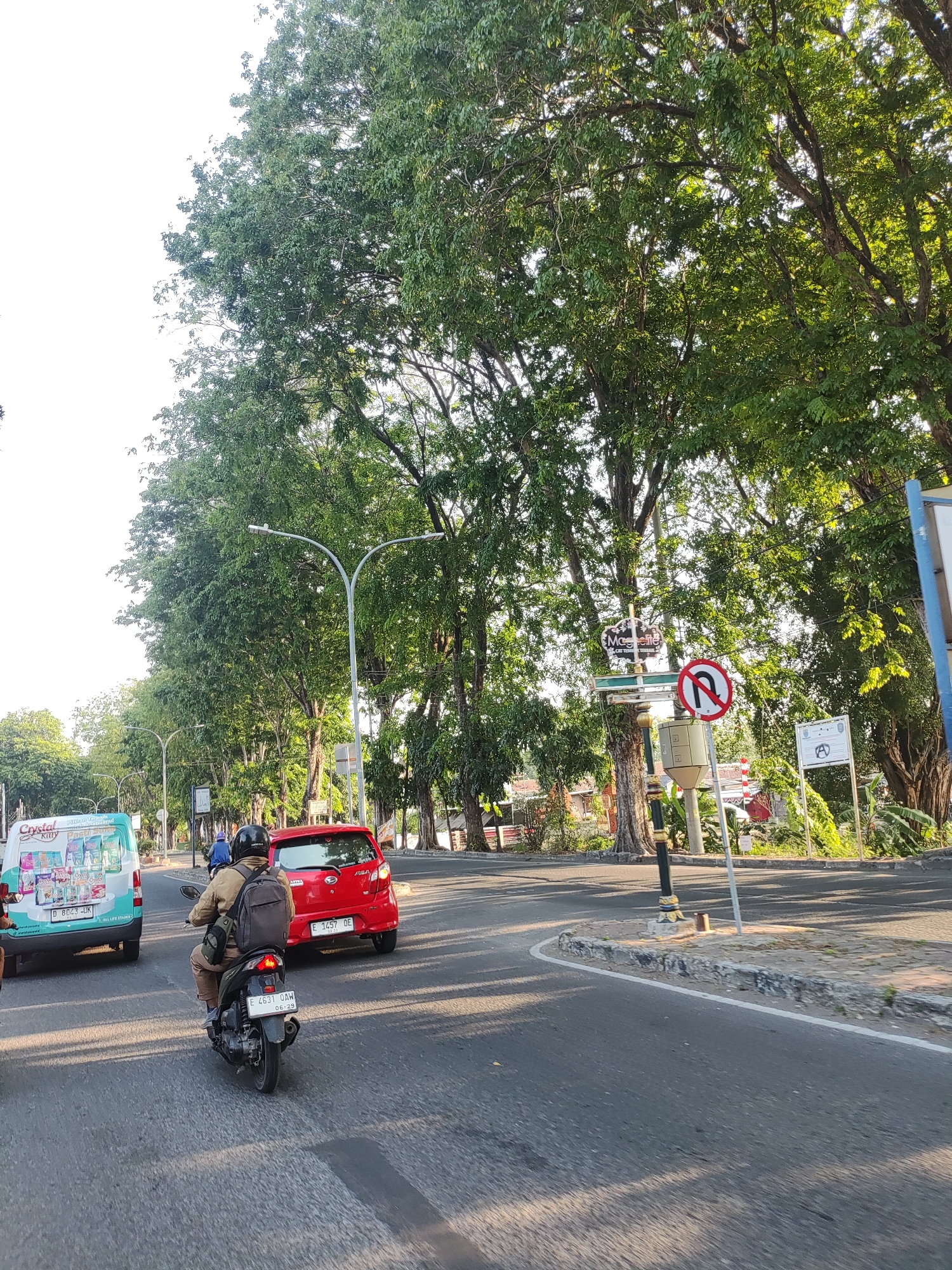 The image captures a street scene in Cirebon, Indonesia, on a sunny day. The perspective is from a vehicle moving on a paved road lined with large trees casting shadows. In the foreground, a black motorcycle with license plate E 4631 DAW is carrying two people. The rider wears a light brown jacket and a black helmet, while the passenger, likely a woman based on visible hair, wears a blue shirt and a black helmet. The motorcycle is in motion, following a red car with license plate E 1457 DAW. To the left, a white van with the branding "Crystal Kitty Fresh Drink" and license plate D 8023 UKR is also moving on the road. A traffic sign prohibiting U-turns is visible on the right side of the road, next to a lamppost. Further back, more trees and buildings suggest an urban environment. The overall atmosphere is one of everyday traffic flow under a bright sky.